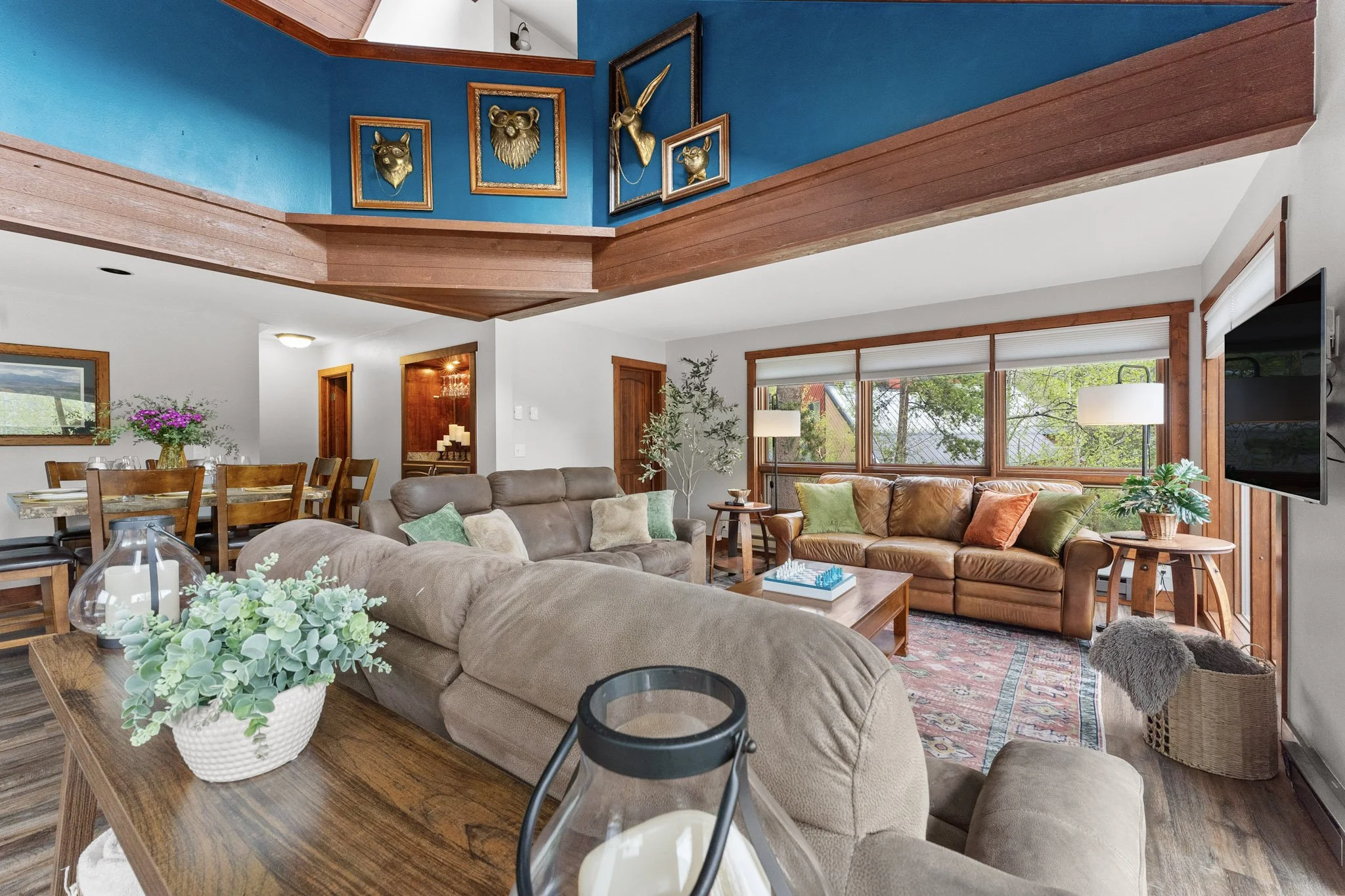 Living room with a brown leather sofa, beige sofa, wooden coffee table, and large windows with blinds showing trees outside. There's a dining area with a wooden table and chairs in the background, decorative plants, and wall art with animal head moun
