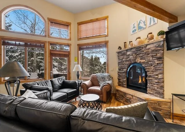 Living room with large windows showing snow-covered trees outside, featuring black leather sofas, a brown armchair with a throw blanket, a stone fireplace with decorative vases on top, and a wall-mounted TV.