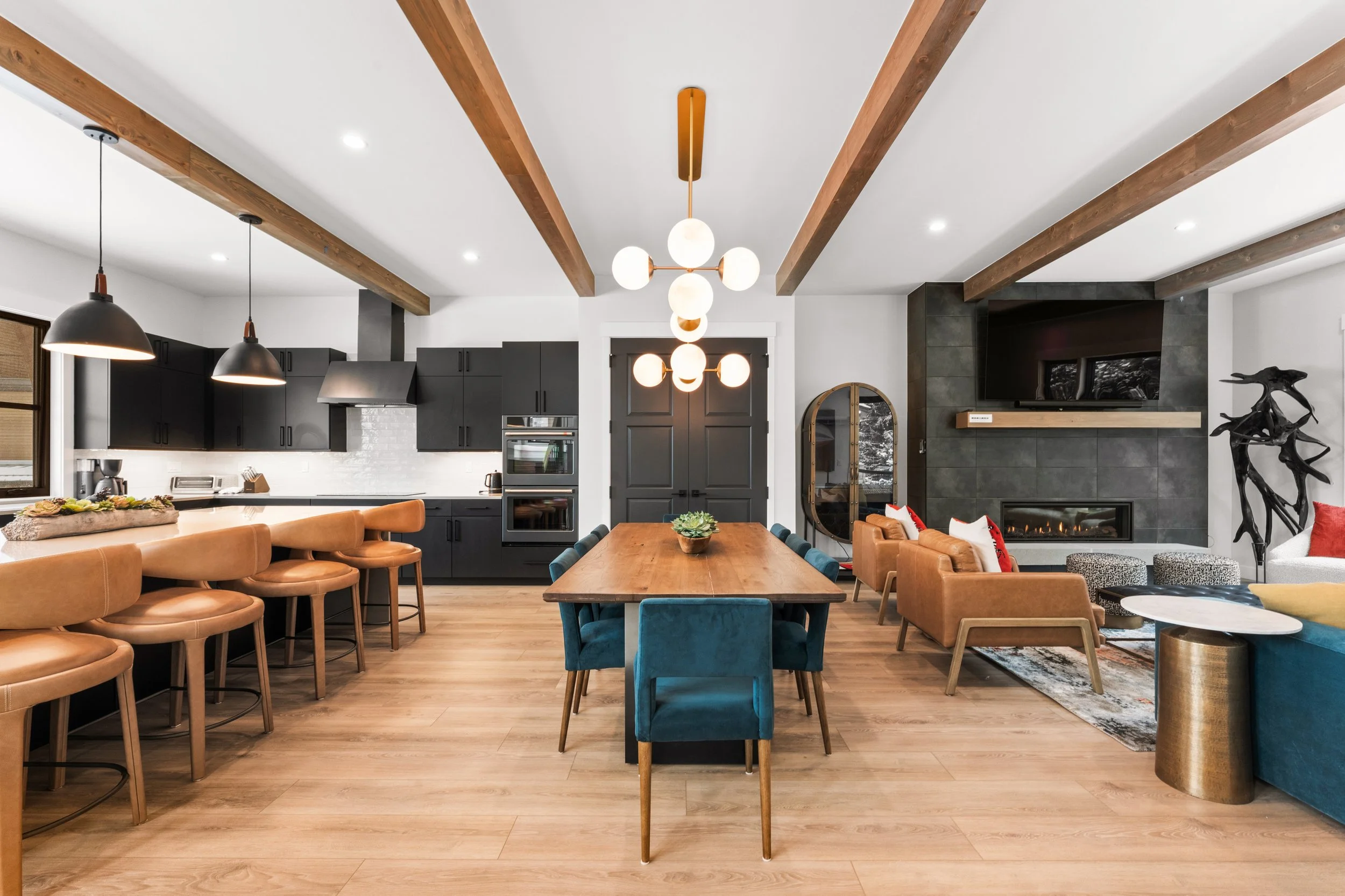 Modern open-concept living and kitchen area with wooden beams, black cabinetry, and a variety of seating including tan, blue, and orange chairs.
