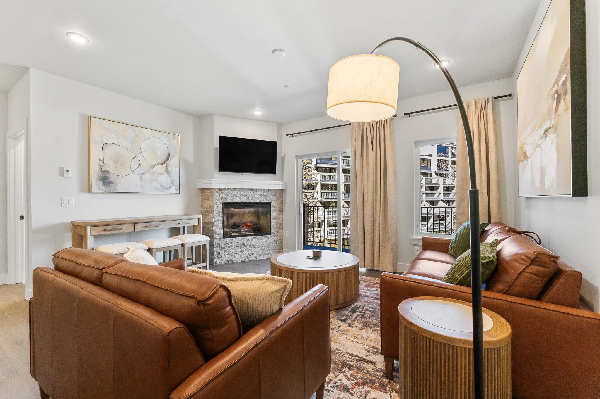 Cozy living room with two brown leather sofas, a white and wood coffee table, fireplace with stone surround, flat-screen TV above fireplace, abstract artwork on white walls, large window with beige curtains, and a standing lamp.