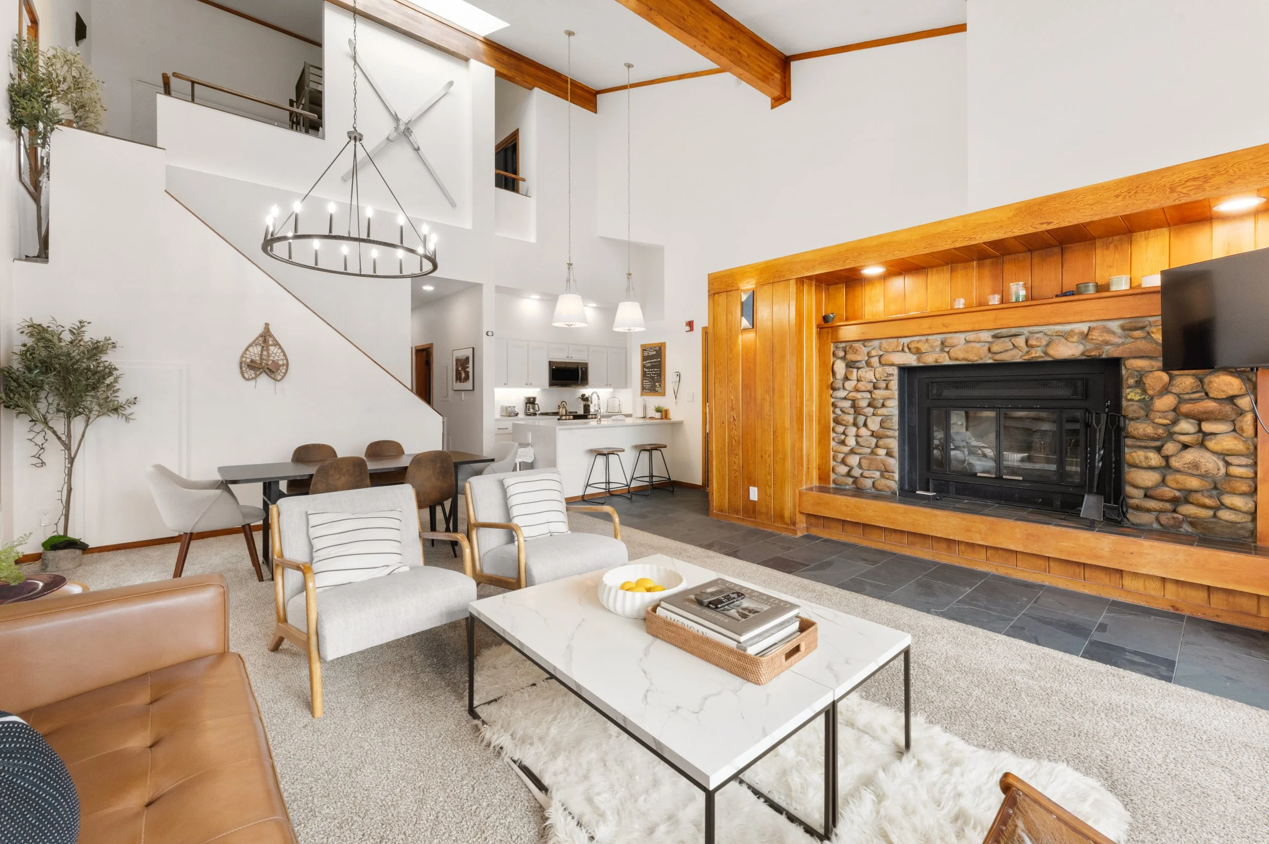 A spacious living room with a high ceiling featuring wood beams and a stone fireplace. The room is decorated with modern furniture, a black chandelier, a large flat-screen TV, and plants, creating a cozy and stylish atmosphere.