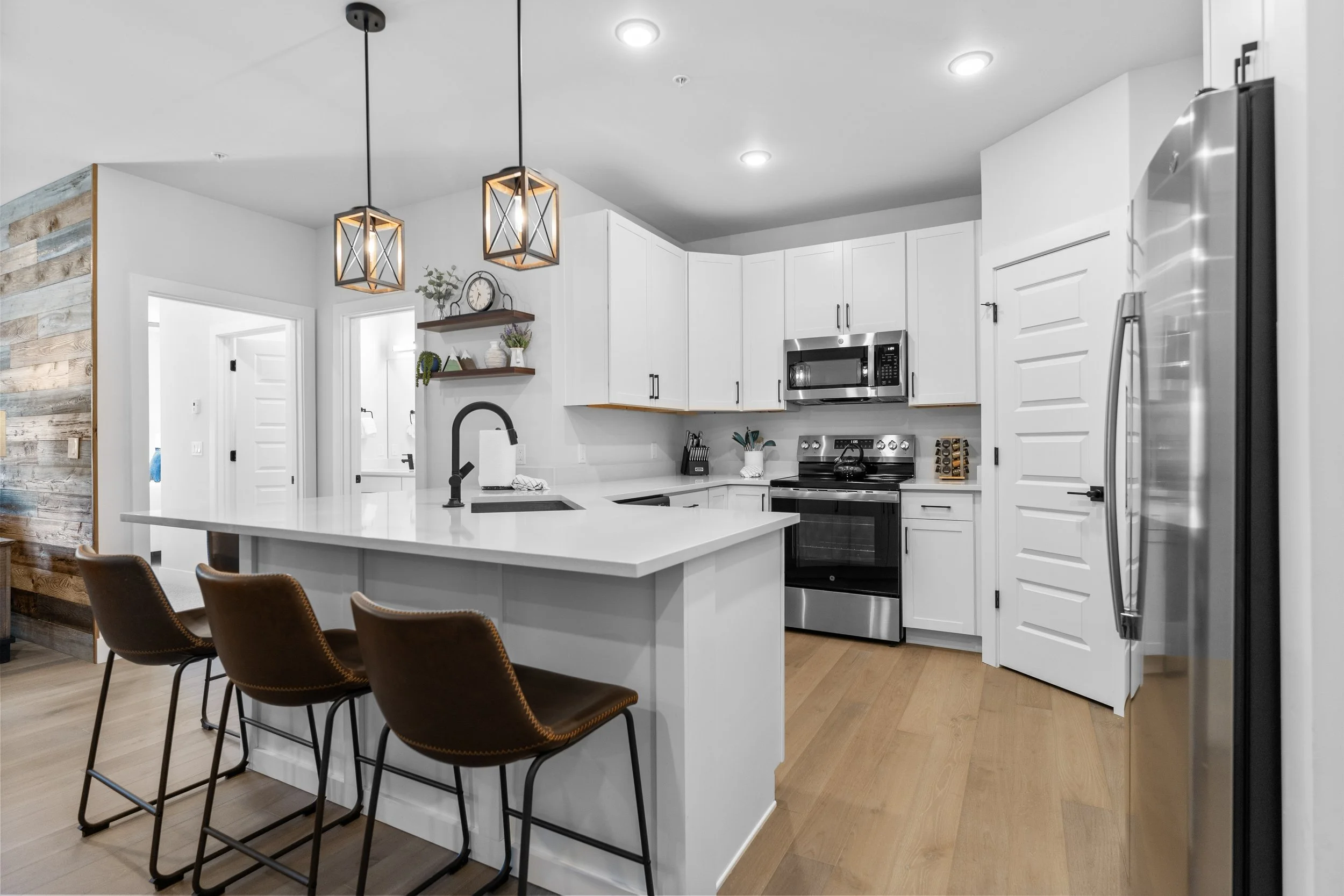 Modern white kitchen with island, black hardware, stainless steel appliances, pendant lights, wooden floor, bar stools, and decorative shelves