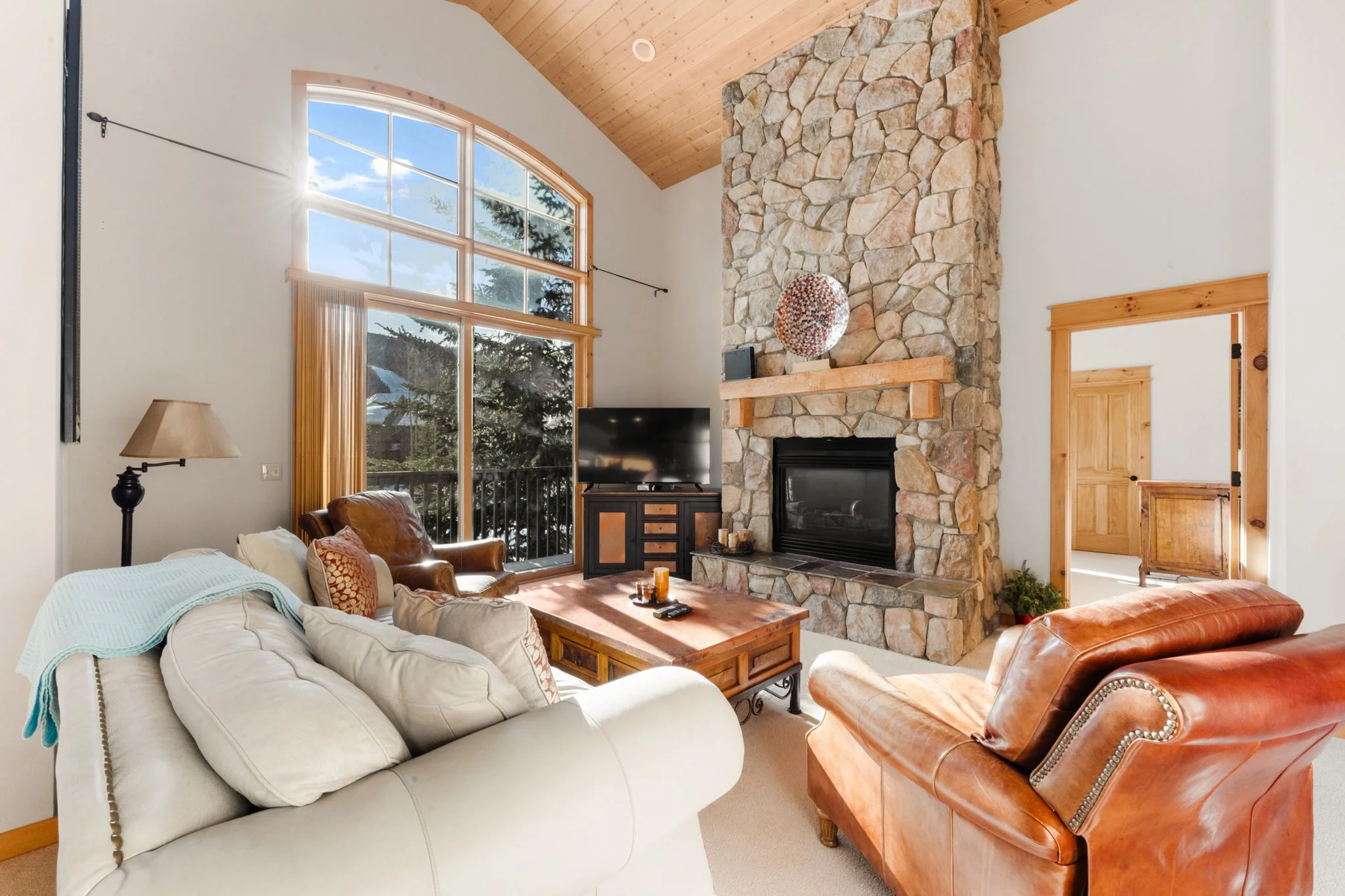 Living room with large arched window, stone fireplace, leather armchair, beige sofa, wooden coffee table, TV, and neutral walls with wooden trim.