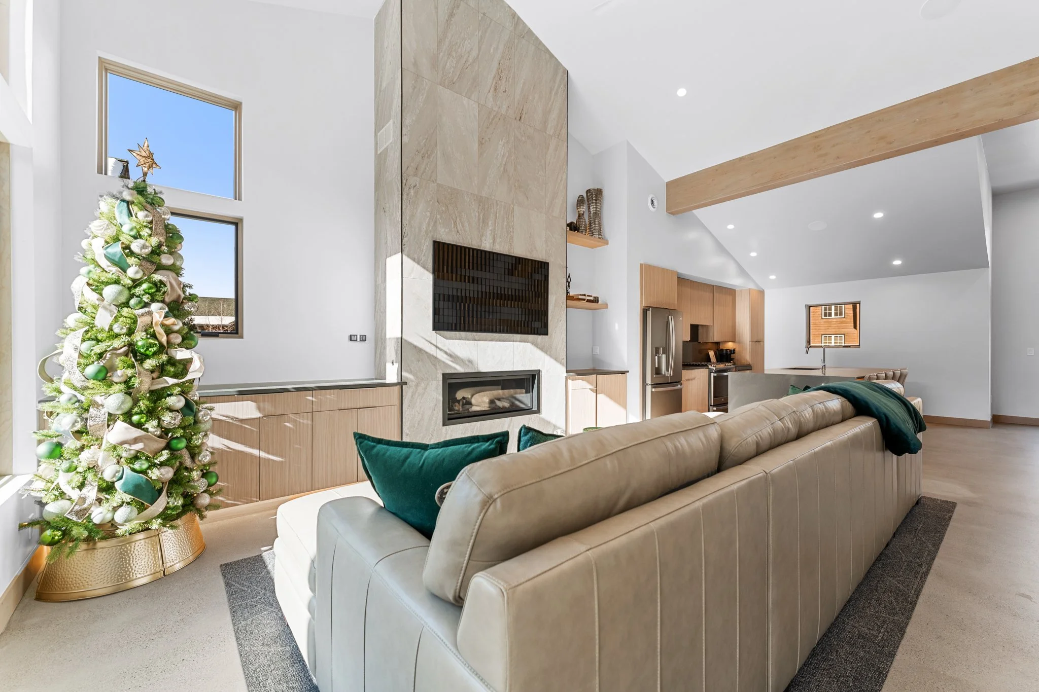 Modern living room with a beige leather sofa, green pillows, a decorated Christmas tree with gold and green ornaments, and a wall-mounted fireplace with a TV above. The room has wooden accents, white walls, large windows, and high ceilings with reces