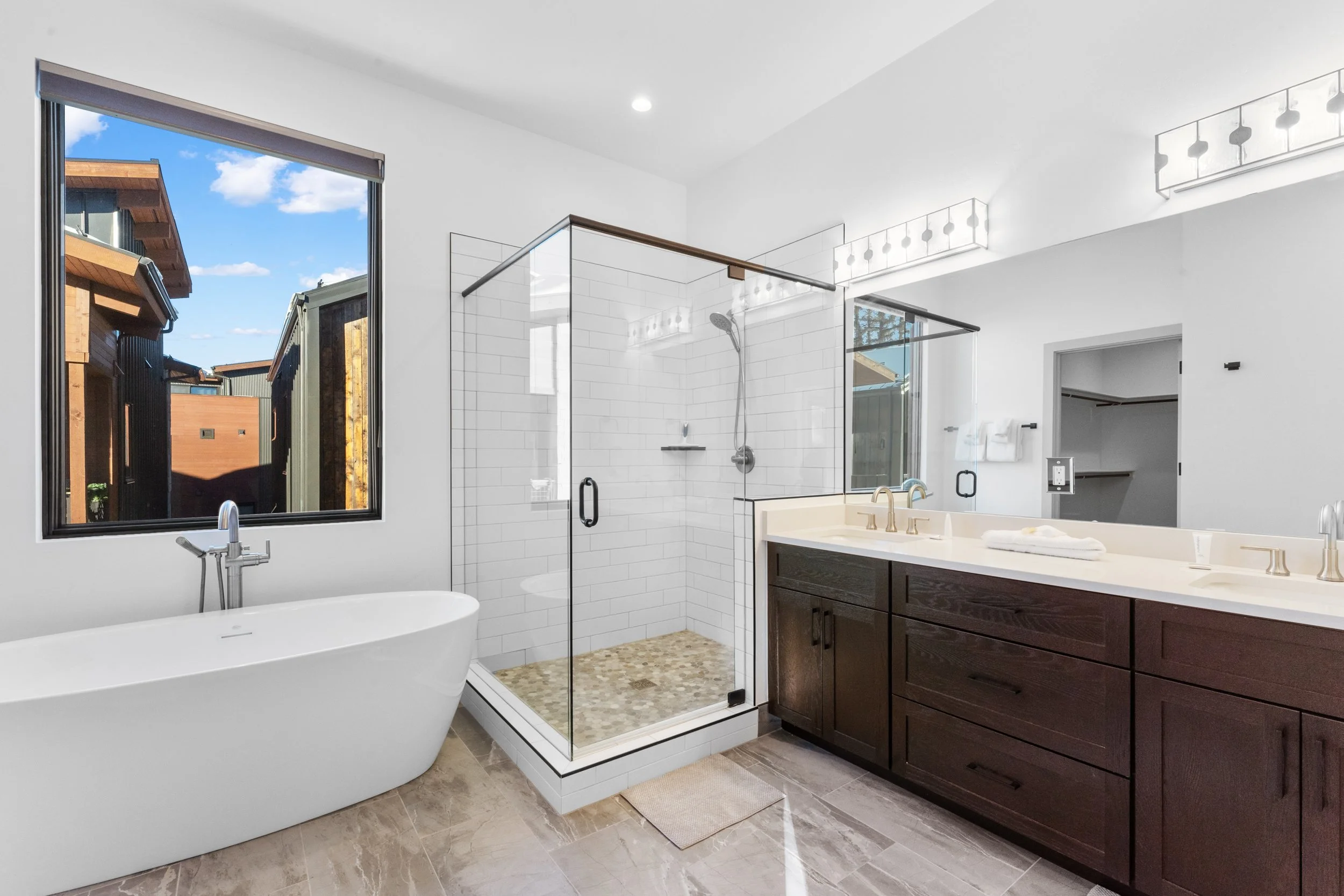 Modern bathroom featuring a bathtub, a glass-enclosed shower with white subway tiles, a dark wood vanity with a white countertop, double sinks, and a large window showing a backyard with a wooden fence and neighboring houses.