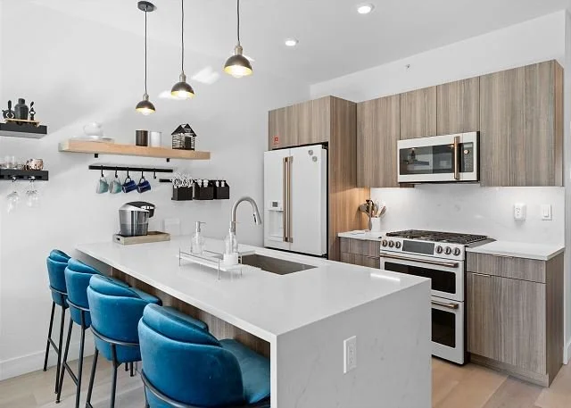 Modern kitchen with white countertops, wooden cabinets, stainless steel appliances, black light fixtures, and blue bar stools.