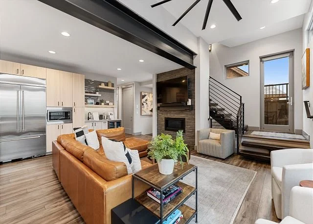 Modern living room with tan leather sofa, white armchair, fireplace, open kitchen, and staircase with metal railing leading to an outdoor deck.