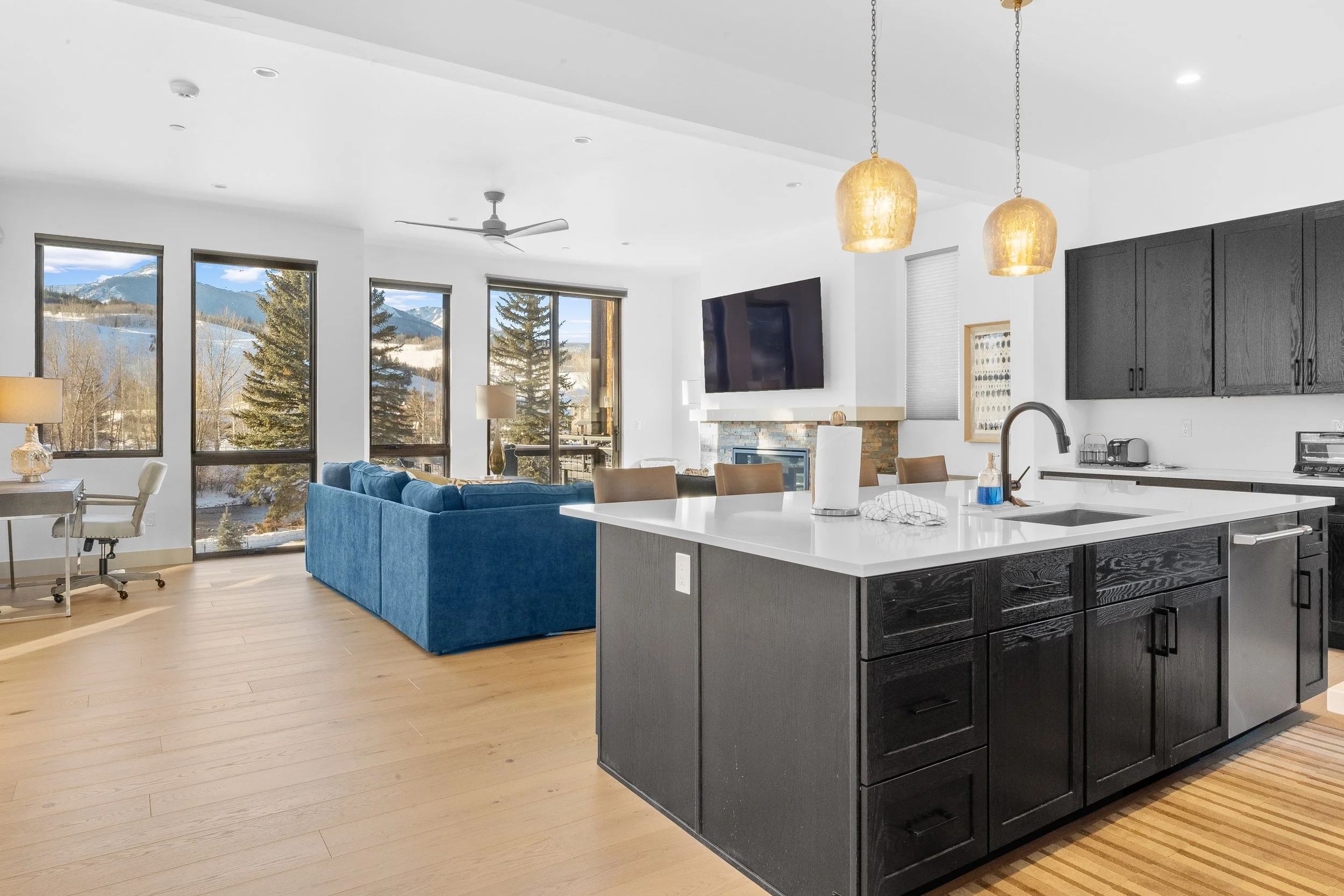 Open-concept living room and kitchen with large windows and mountain view, black kitchen cabinets, white countertops, blue sofa, wood flooring, and pendant lighting.