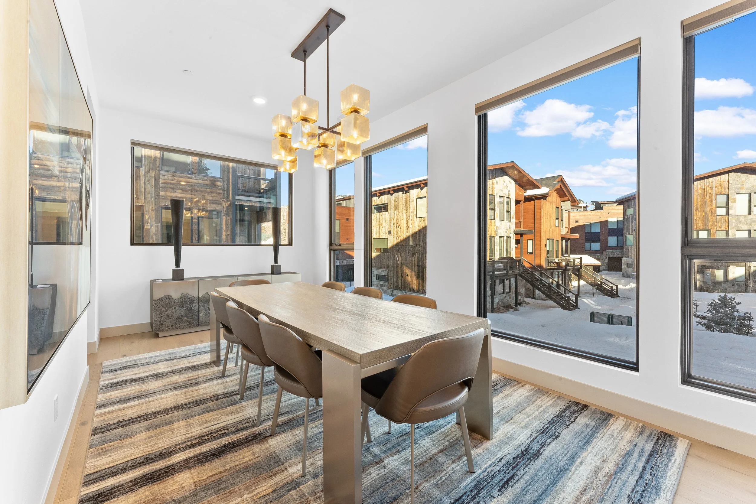 Modern dining room with a wooden table, six beige chairs, large windows showing snowy outdoor buildings, a chandelier with square glass shades, and a patterned area rug.