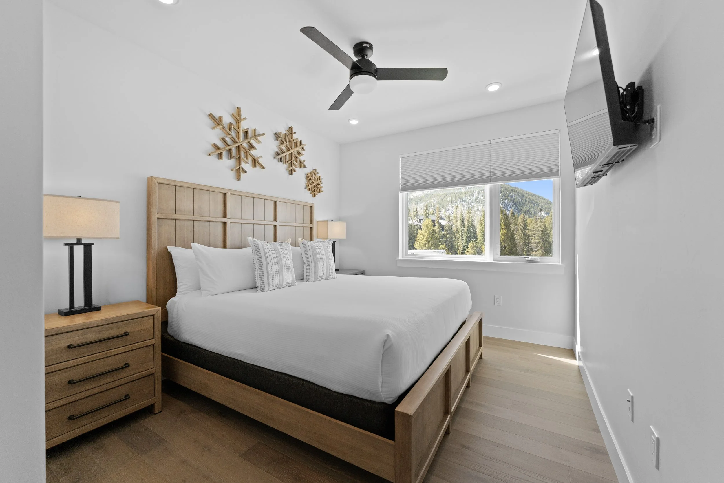 Modern bedroom with white walls, wooden bed frame, white bedding, three decorative pillows, wooden nightstand with lamp, window with view of trees and mountains, mounted TV, ceiling fan, and snowflake wall decorations.