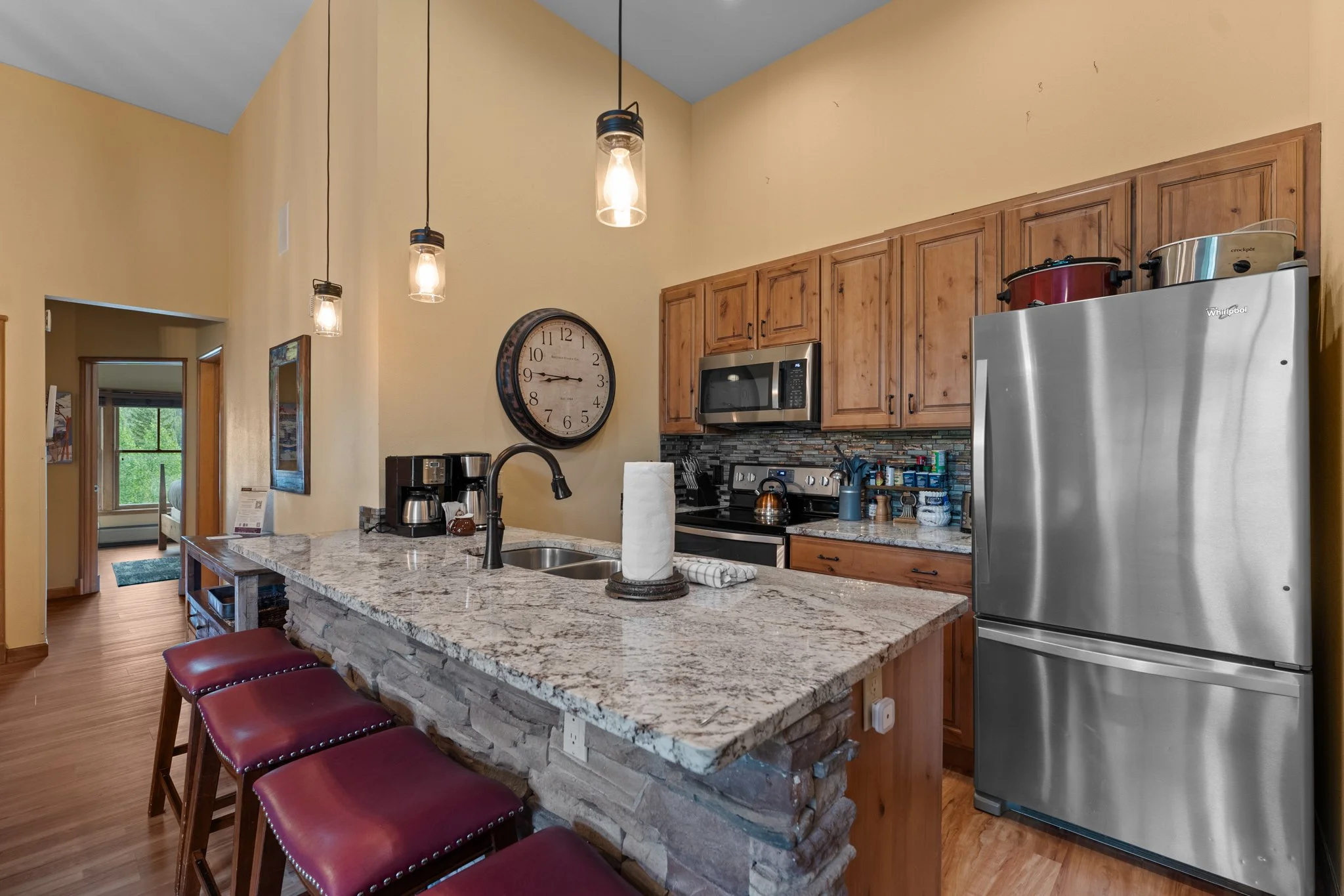 Kitchen with wooden cabinets, granite countertop island, stainless steel refrigerator, microwave, coffee maker, and three burgundy bar stools.