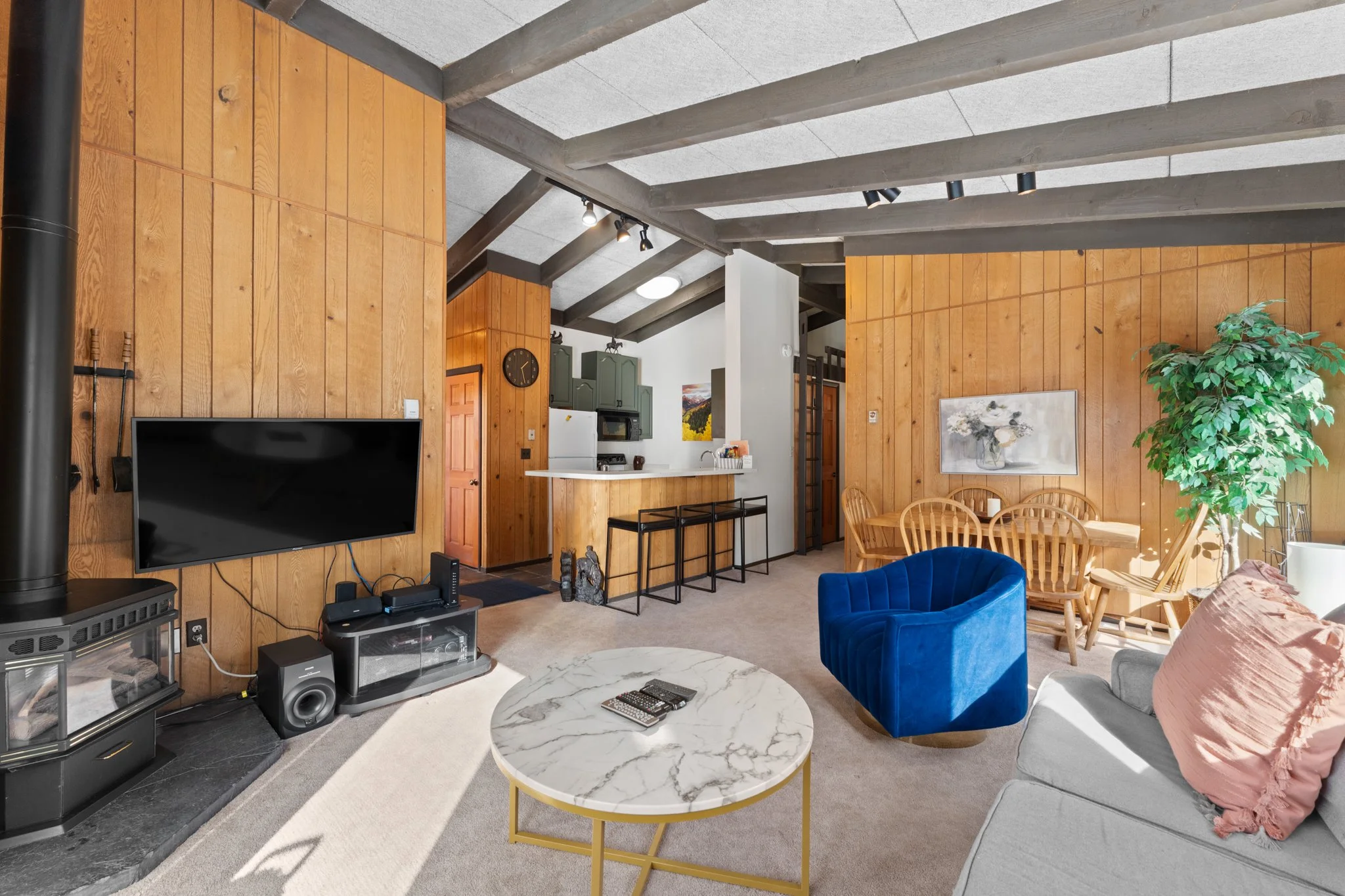 Living room with a wood-paneled wall, a white ceiling with exposed beams, a flat-screen TV, a marble coffee table, a blue velvet chair, a beige sofa with pink pillows, a large potted plant, and a wooden dining table with chairs.