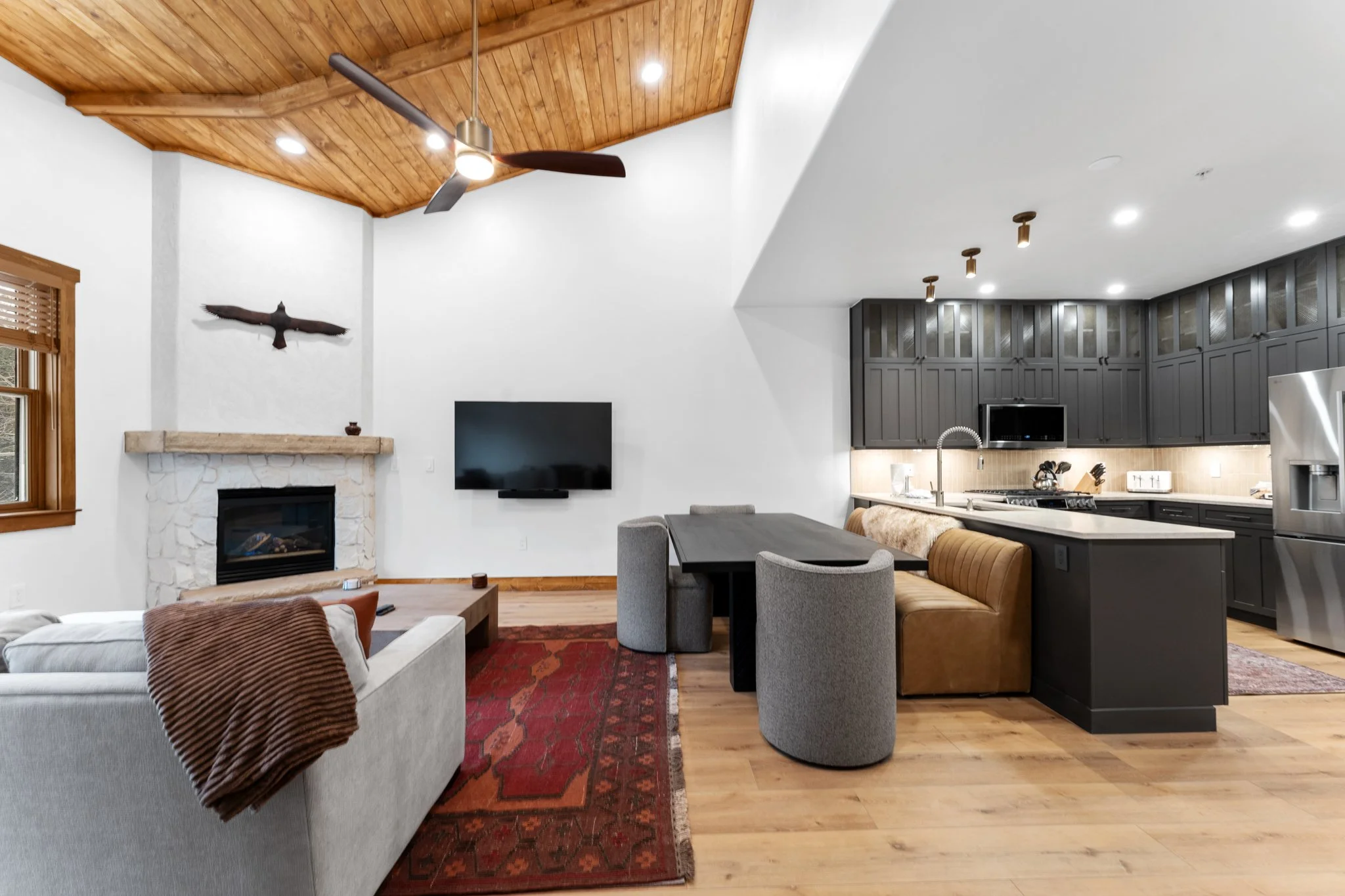 Interior view of a living room and kitchen with wood ceiling, fireplace, wall-mounted TV, gray and brown furniture, black kitchen cabinets, stainless steel refrigerator, and wood flooring.