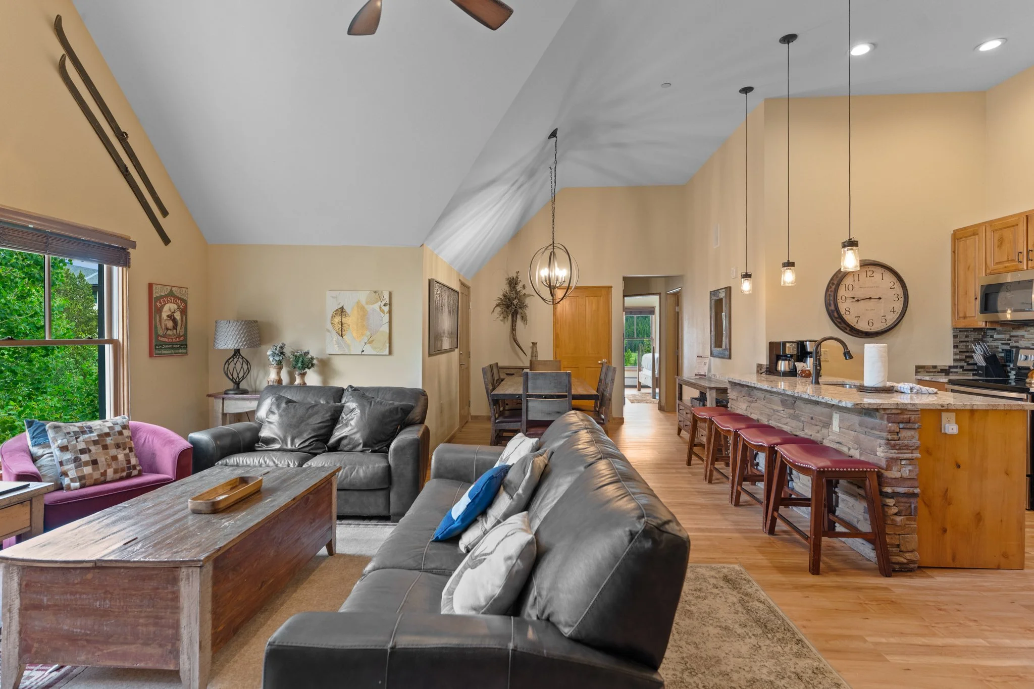 Open-concept living room and kitchen with high vaulted ceilings, hardwood floors, gray leather sofa, pink armchair, wooden coffee table, dining table with six chairs, kitchen bar with three stools, and large window with green trees outside.