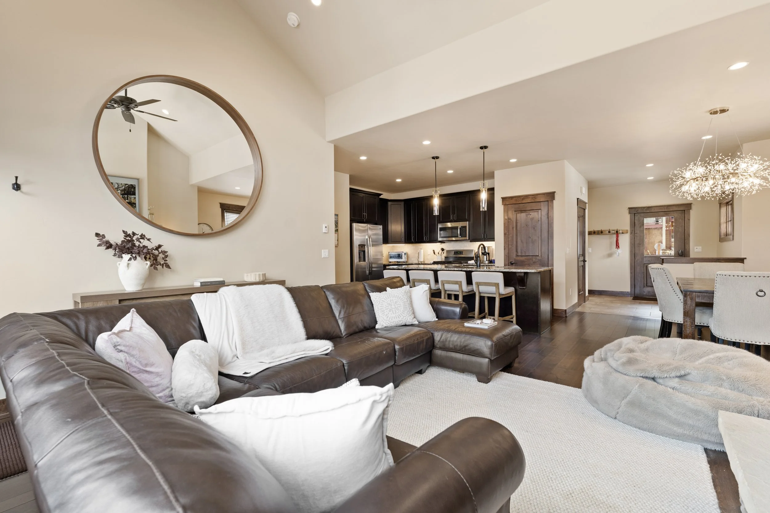 Open-concept living room and kitchen with beige walls, a large leather sectional sofa with white and light-colored pillows, a wall mirror, a wooden sideboard with a vase, dark wood flooring, and pendant lights over the kitchen island.