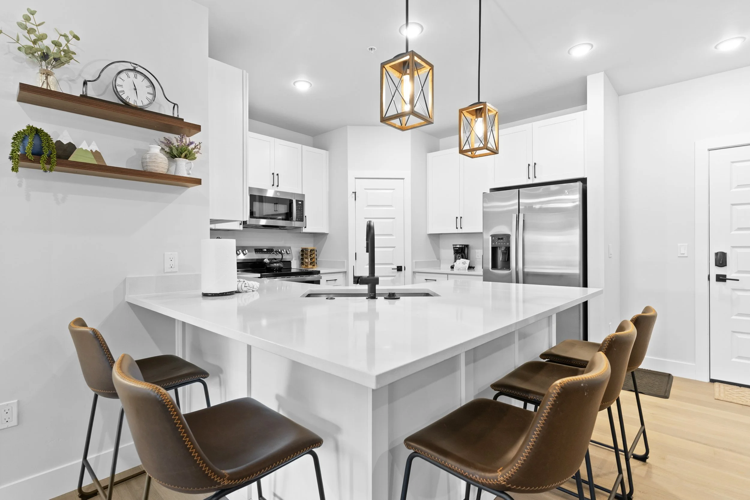 Modern white kitchen with a large breakfast bar, brown leather stools, white cabinetry, and stainless steel appliances. Two pendant lights hang above the bar. Shelves with decor on the wall.