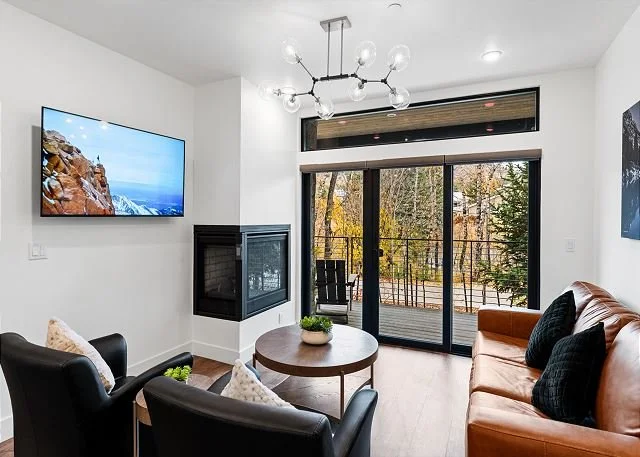 Modern living room with fireplace, round coffee table, leather sofa, armchairs, wall-mounted TV, large glass sliding door leading to a deck, and view of trees outside.
