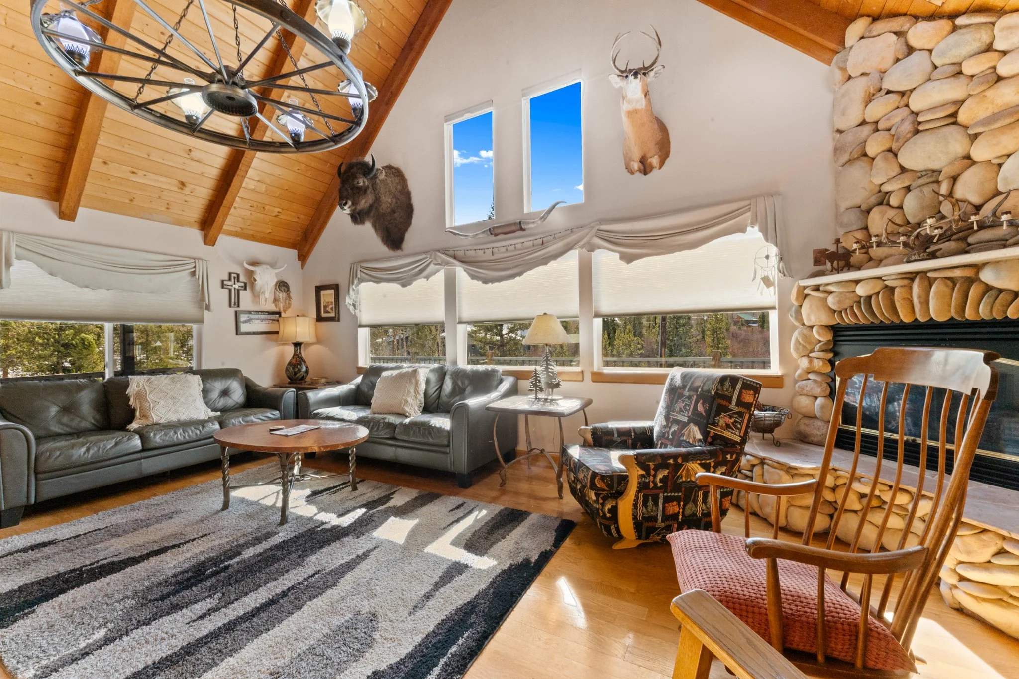 Living room with large windows, wooden ceiling, mounted animal heads including a deer and a bison, a stone fireplace, comfortable sofas, and wooden chairs.