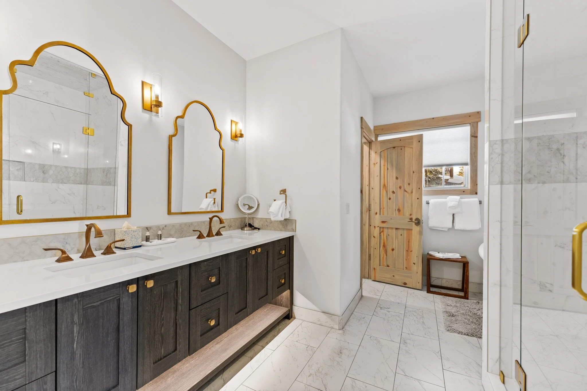 Bathroom with double vanity sinks, dark wood cabinets, gold fixtures, two mirrors, and white marble countertops. There are wall-mounted light fixtures, a tissue box, a small clock, and towel holders. The background includes a wooden door leading to a
