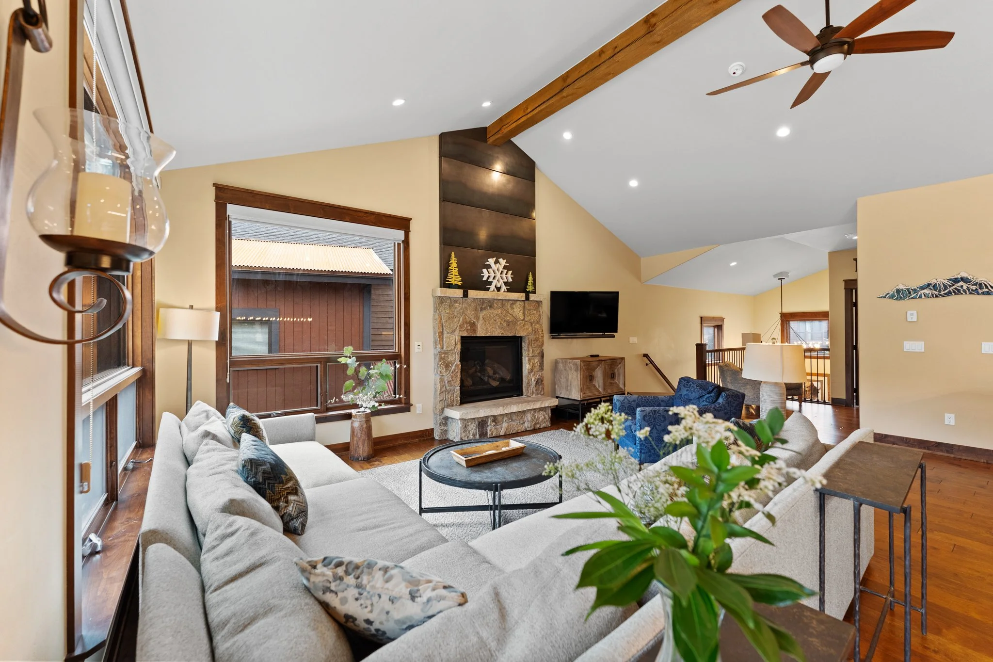 Living room with a beige sofa, blue armchair, stone fireplace, wall-mounted TV, wooden accents, and large windows letting in natural light.