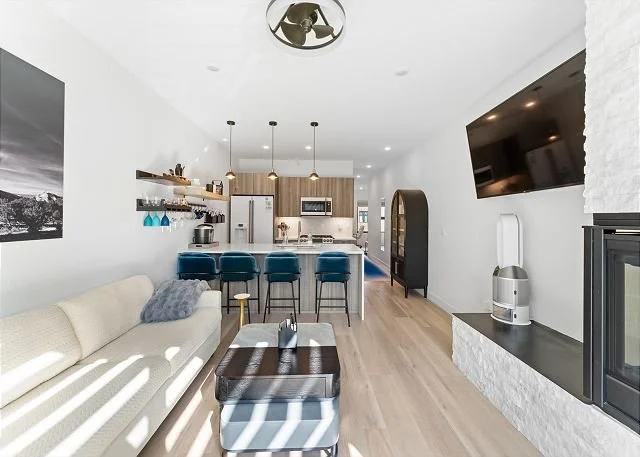 Open-concept living room and kitchen with white walls, light wood floors, a white sofa with a gray pillow, a wooden coffee table, blue bar stools at a kitchen island, and a flat-screen TV on the wall.