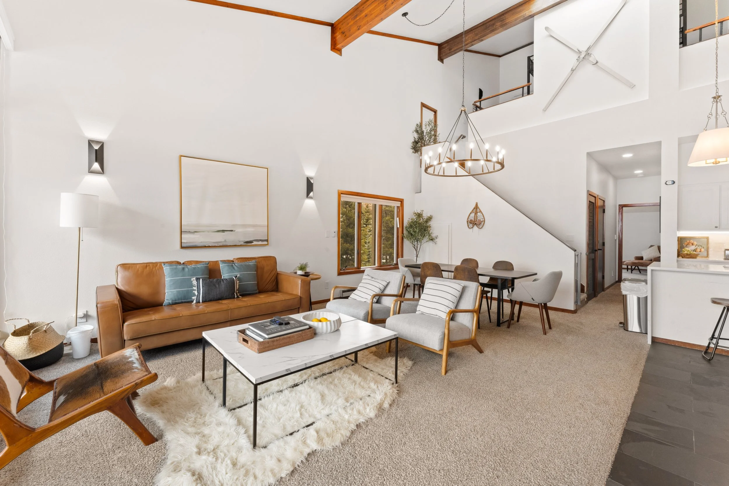 A spacious living room with high vaulted ceilings, white walls, and exposed wooden beams. It features a brown leather sofa with pillows, two armchairs, a marble-top coffee table, and a shaggy white rug. There are wall-mounted lights, large windows, a