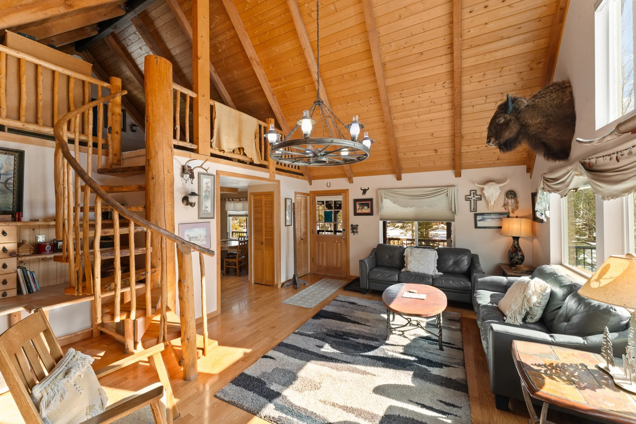 Living room with wooden vaulted ceiling, leather sofas, a patterned rug, large windows, wall decorations including a mounted bison head and a cross, and a chandelier hanging from the ceiling.