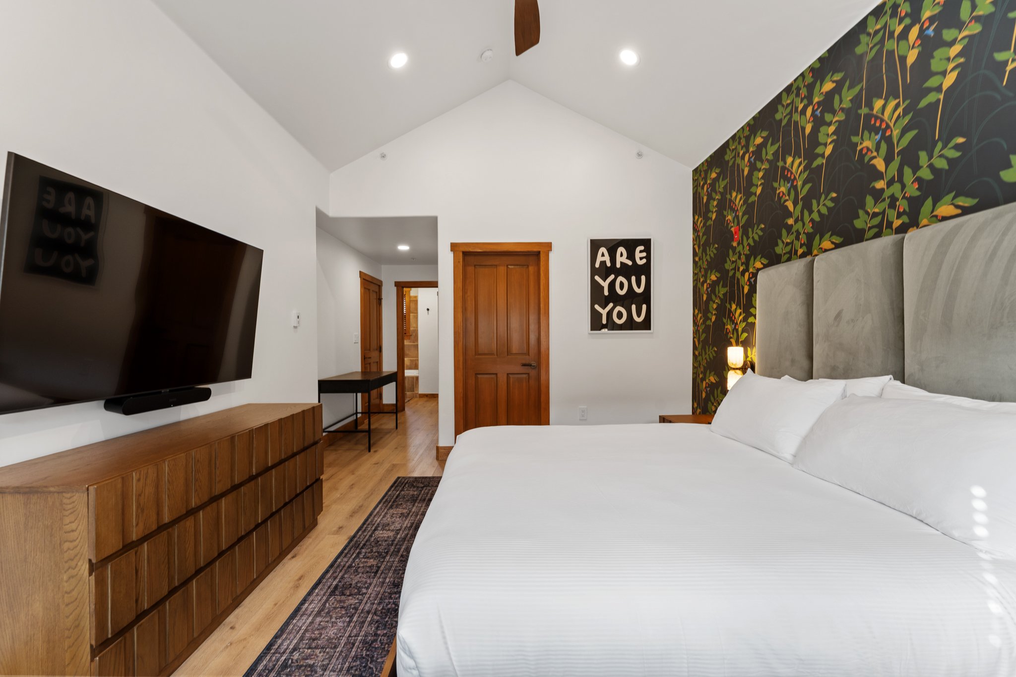 Well-lit bedroom with a king-sized bed, large gray upholstered headboard, dark wallpaper with green leaves and red berries, white walls, wooden dresser, flat-screen TV, and a black framed artwork with the text 'ARE YOU YOU' on the wall.