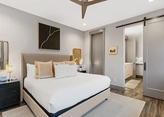 A modern bedroom with a beige upholstered bed, white bedding, and multiple pillows. There are black nightstands with lamps on either side of the bed, a gold abstract wall art above, and a sliding barn door leading to a bathroom with white vanity and 