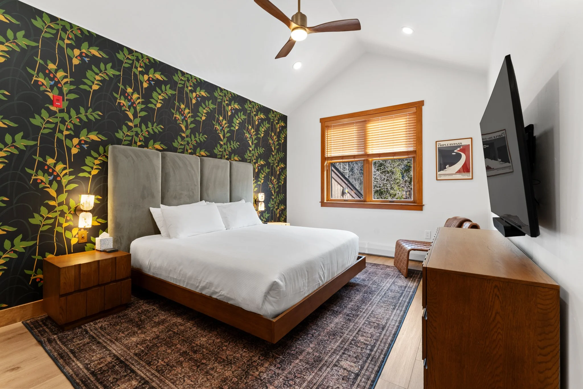 Hotel bedroom with a large bed, wooden nightstand, mounted TV, colorful wallpaper on one wall, window with wooden blinds, ceiling fan, and artwork on the wall.