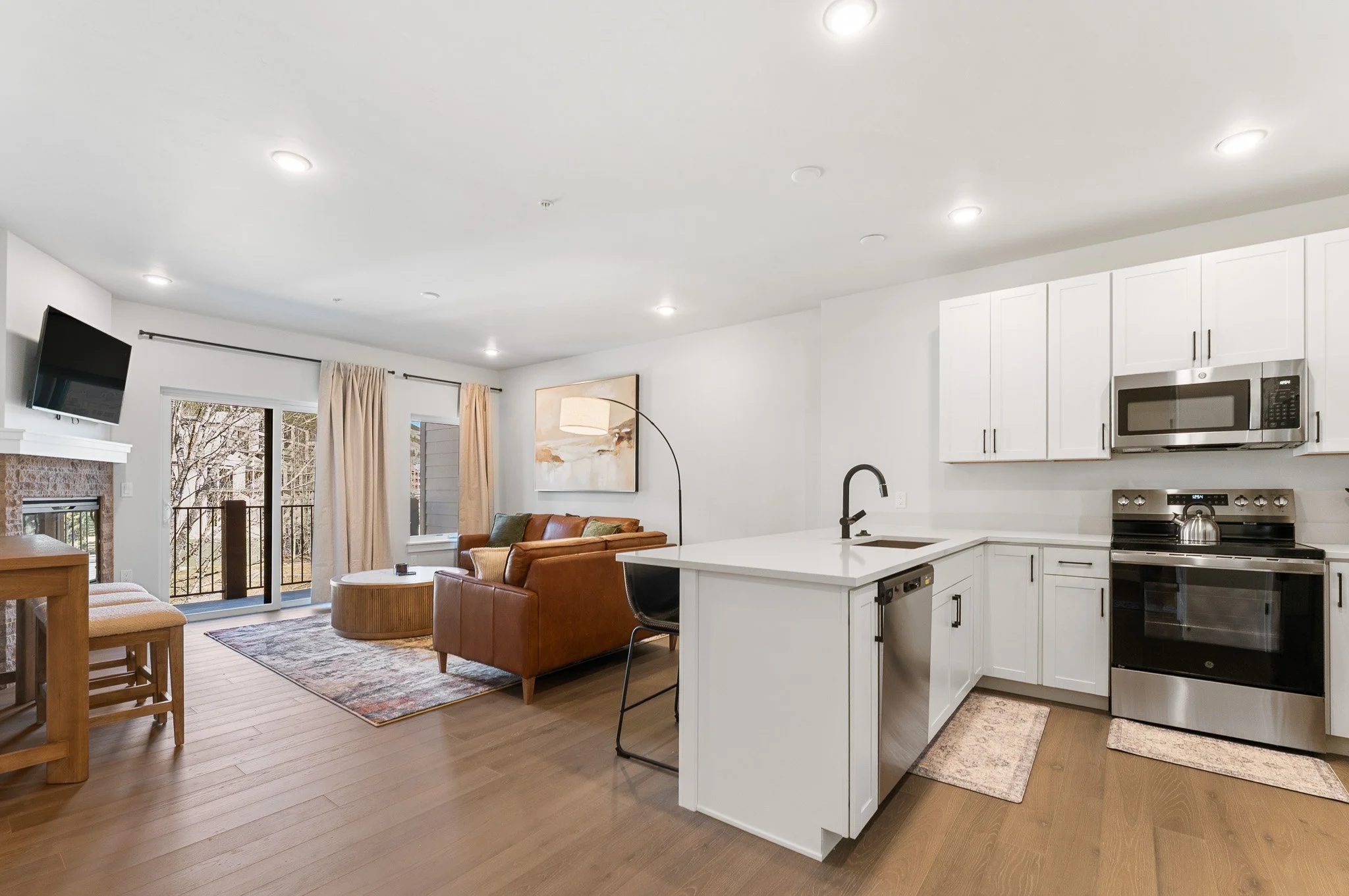 Open-concept living room and kitchen with white cabinets, stainless steel appliances, a leather sofa, a glass sliding door leading to an outdoor balcony, and natural light.