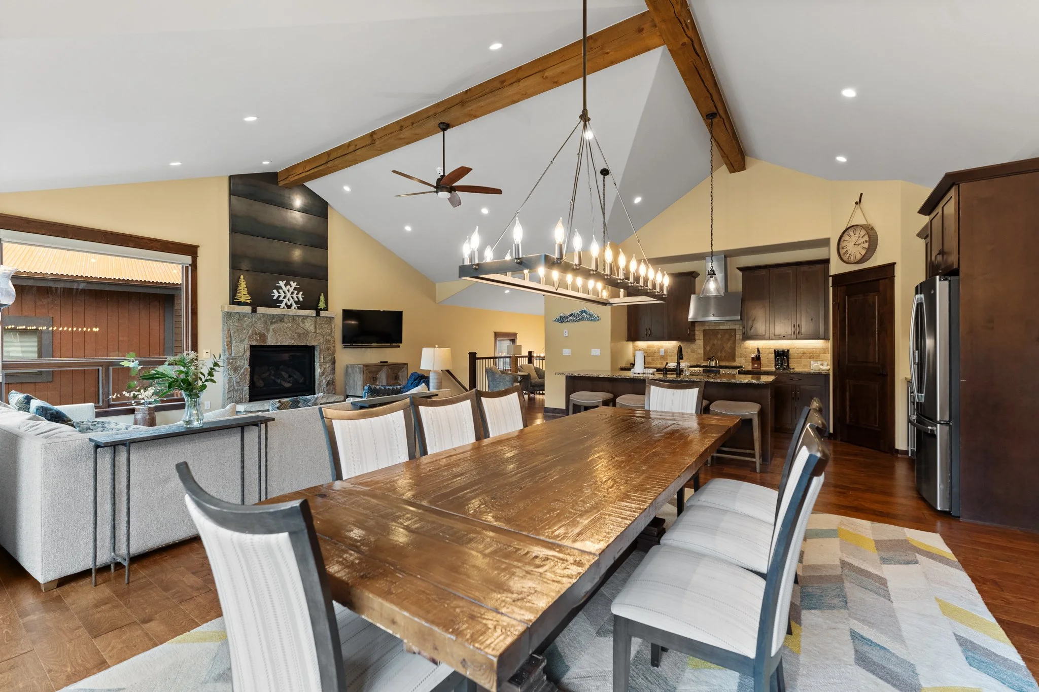 Open-concept living and dining area with a wooden dining table, beige chairs, a kitchen with dark wood cabinets, and a kitchen island. A vaulted ceiling with exposed wooden beams, a chandelier, a ceiling fan, and a stone fireplace in the background.
