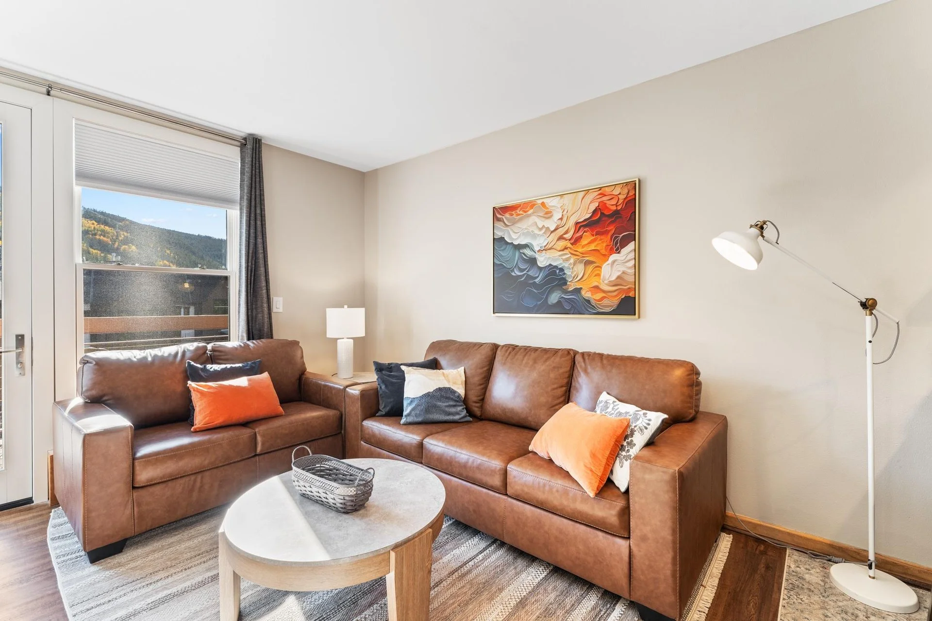 Living room with two brown leather sofas, an abstract painting on the wall, a white floor lamp, a small round wooden coffee table, a window with a view of mountains, and various throw pillows.
