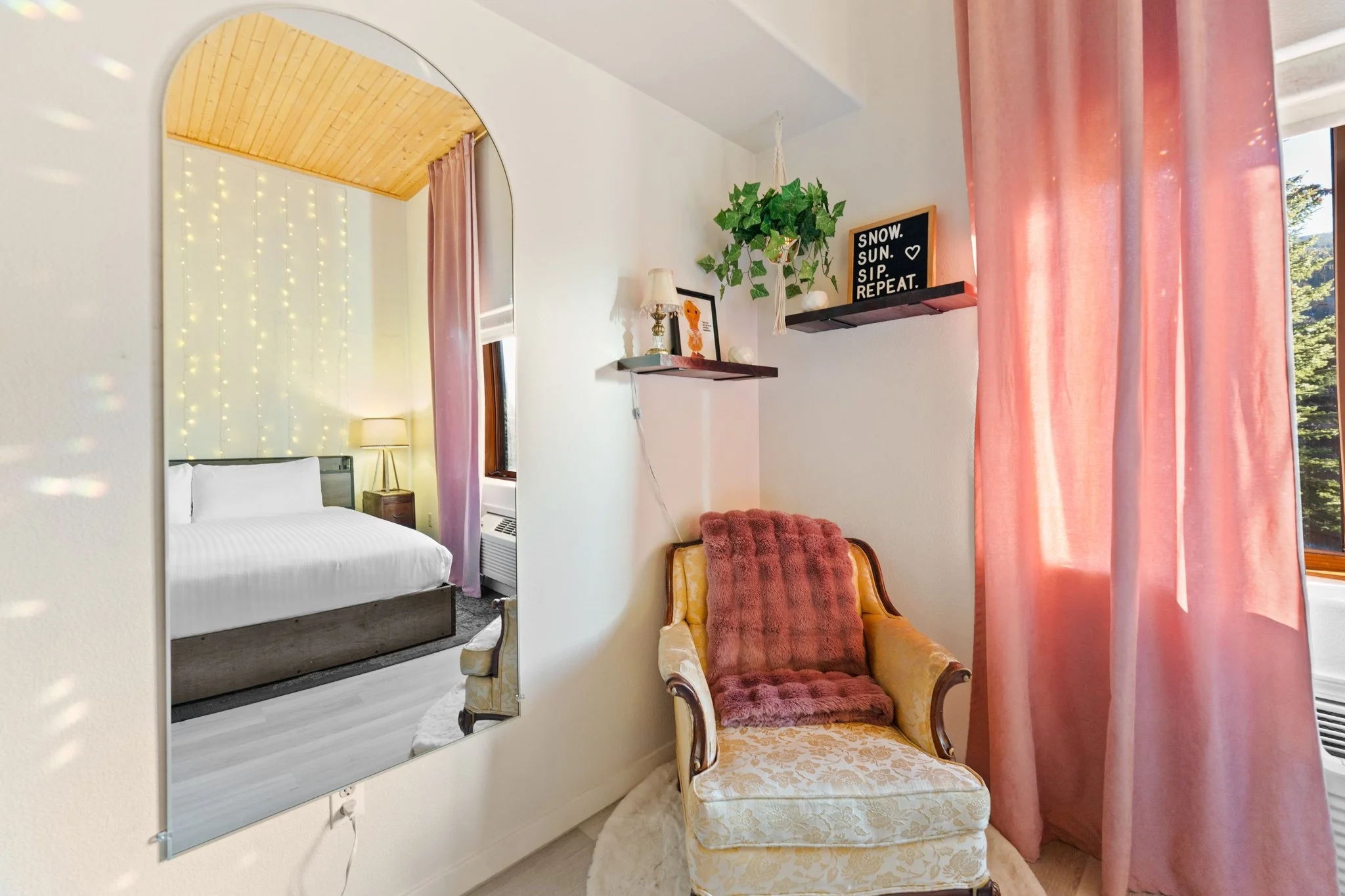 A cozy room with a vintage armchair, pink curtains, framed art and a mirror reflecting a bedroom with fairy lights and a bed.