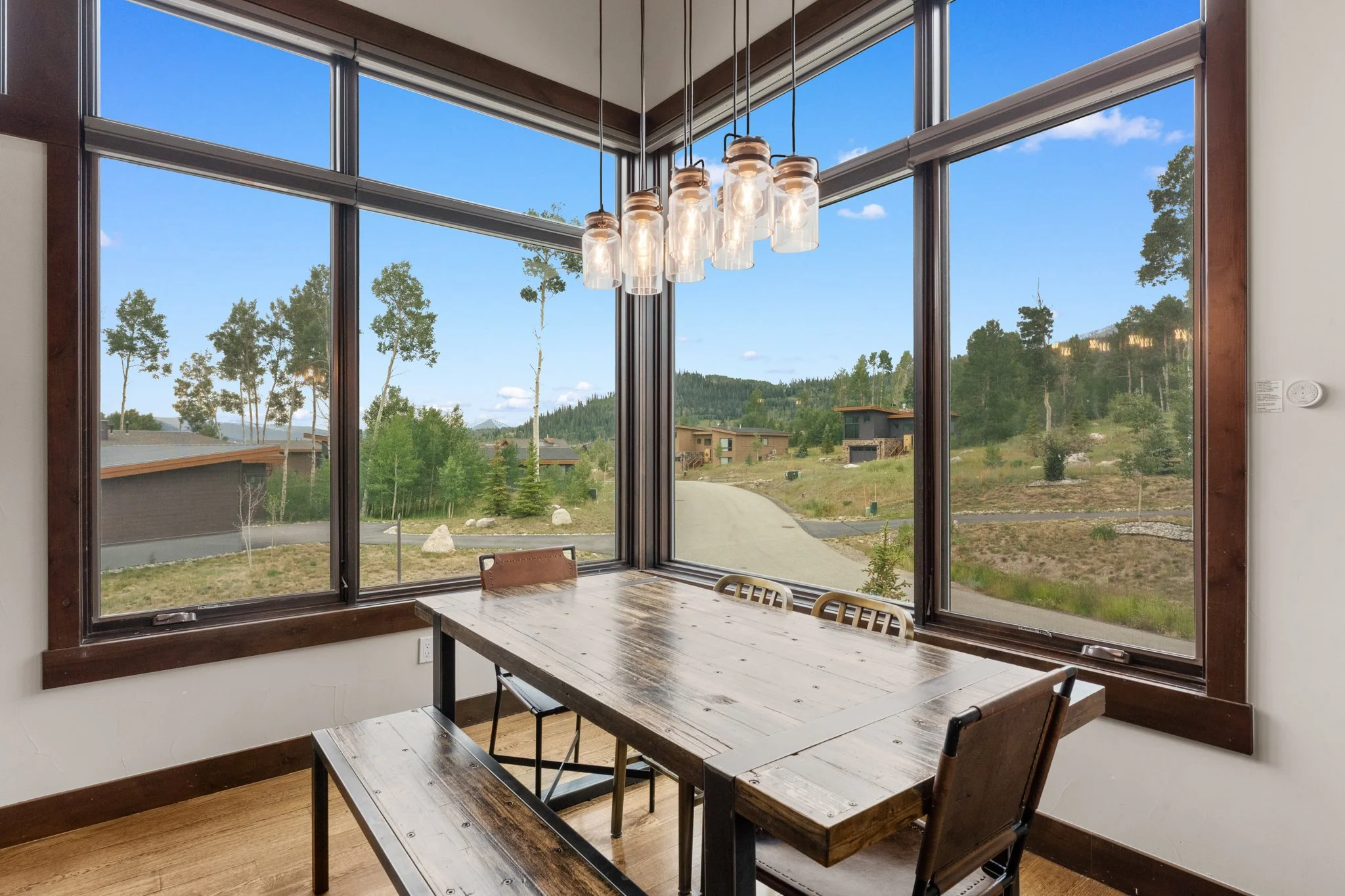 Dining area with large windows showing a scenic outdoor view of trees and houses, wooden table with bench and chairs, modern pendant lighting fixtures