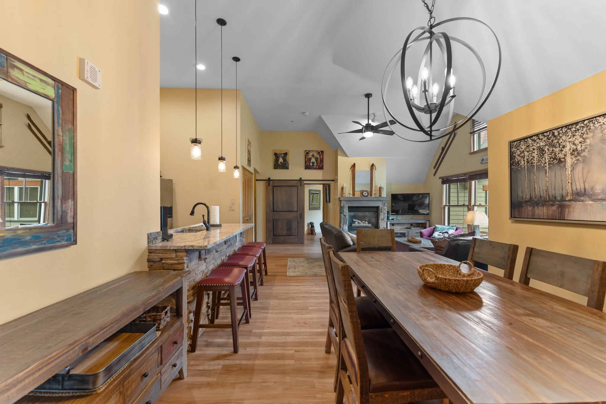 Open-concept living room and kitchen with wooden furniture, a stone fireplace, a large wall art of trees, and a modern chandelier, basked in natural light with hardwood flooring.