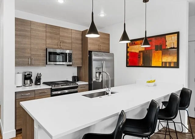 Modern kitchen with wooden cabinets, stainless steel refrigerator, microwave, and coffee maker, black bar stools at a white kitchen island, and abstract colorful painting on white wall.
