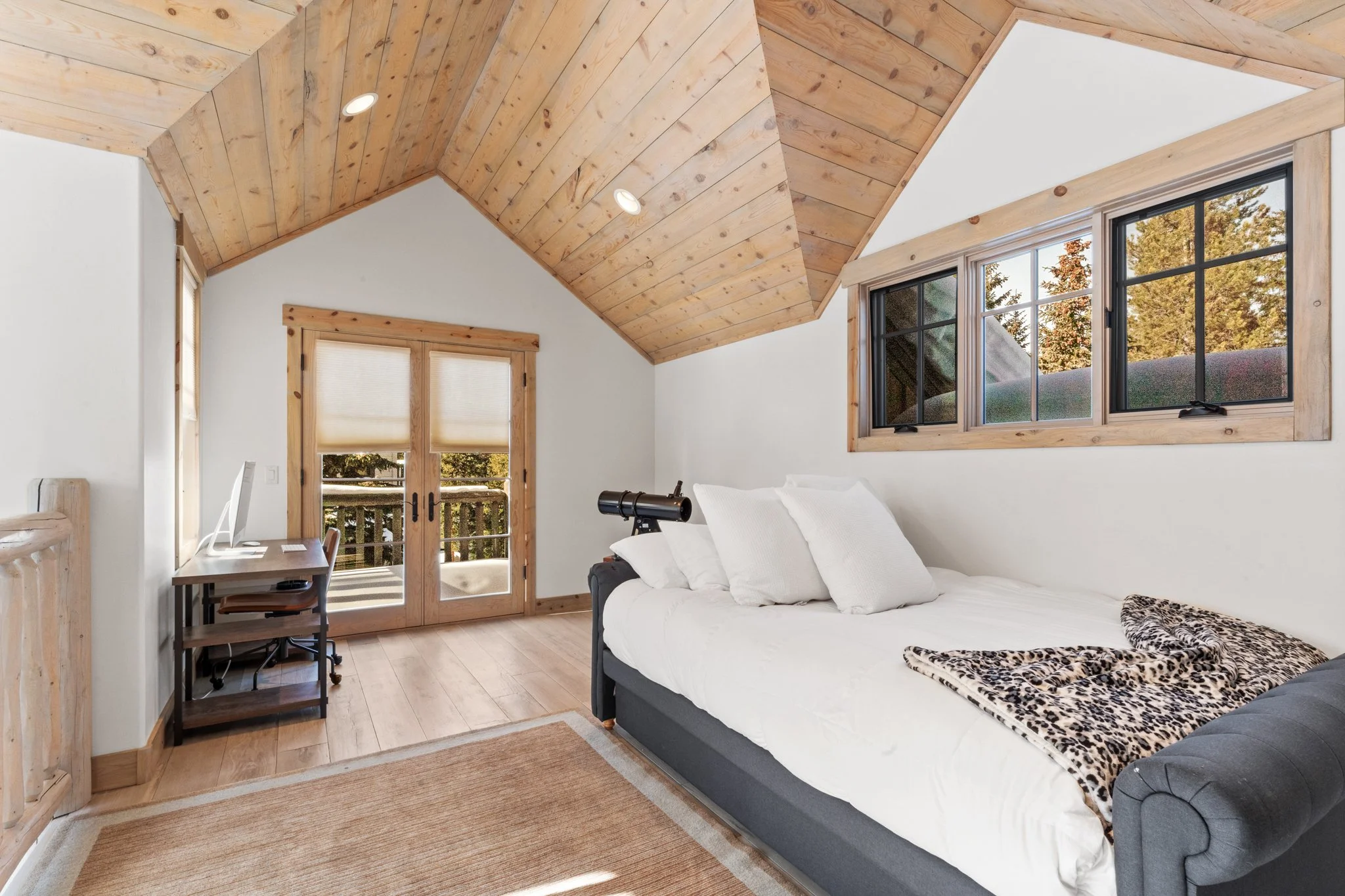 A cozy bedroom with a sloped ceiling made of wood planks, a large bed with white bedding and leopard print blanket, wood-framed windows showing trees outside, a small desk with a computer, and a door leading to a balcony.
