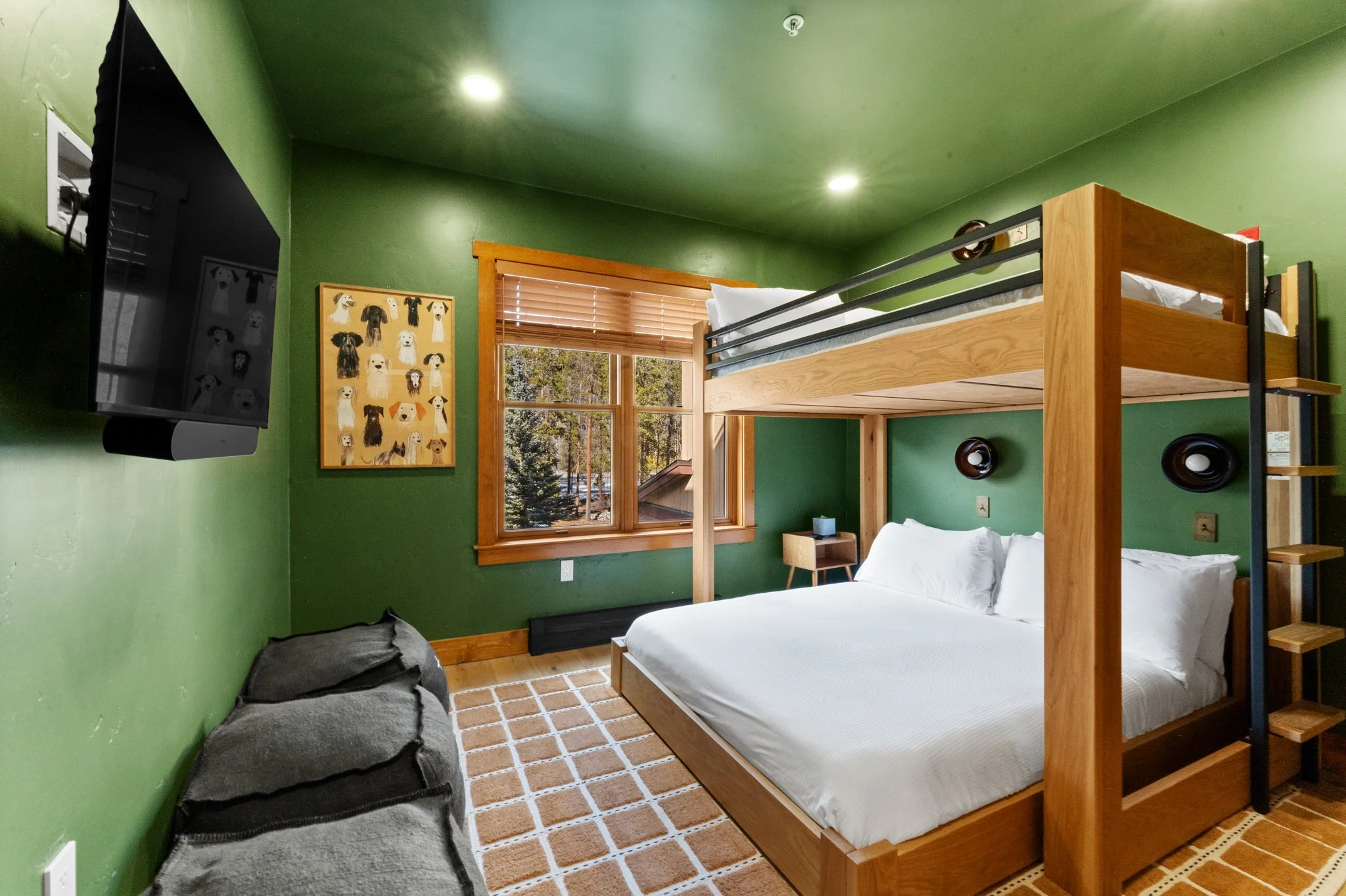 Bedroom with green walls, bunk beds with stairs, window with blinds showing trees outside, wall-mounted TV, dog art picture, bean bag chair, and a beige patterned rug.