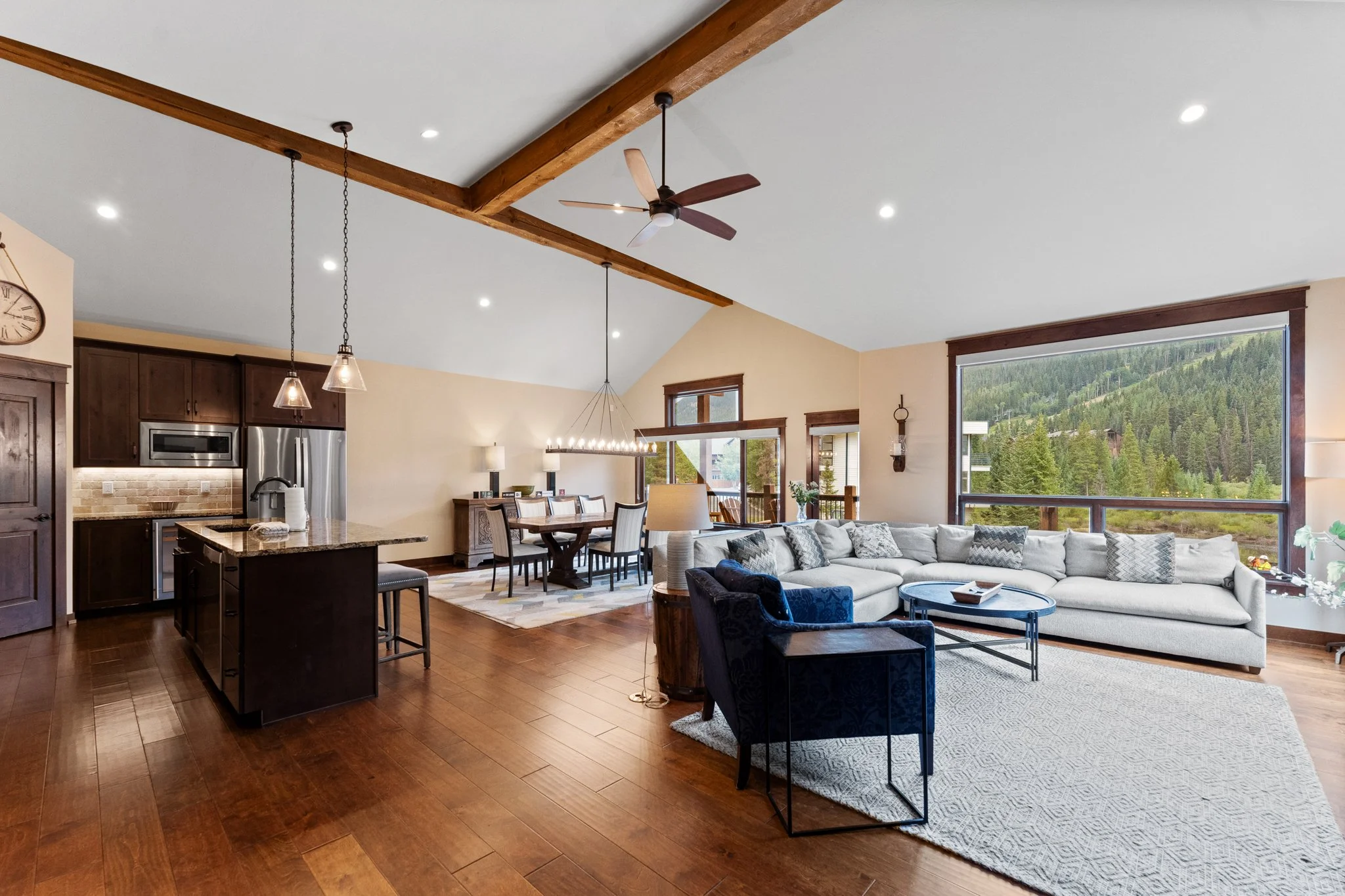 Open-concept living room and kitchen with hardwood floors, large window showing green hills, beige walls, and wooden beams on ceiling. Furniture includes a white sectional sofa, blue armchair, dining table with chairs, and kitchen island with barstoo