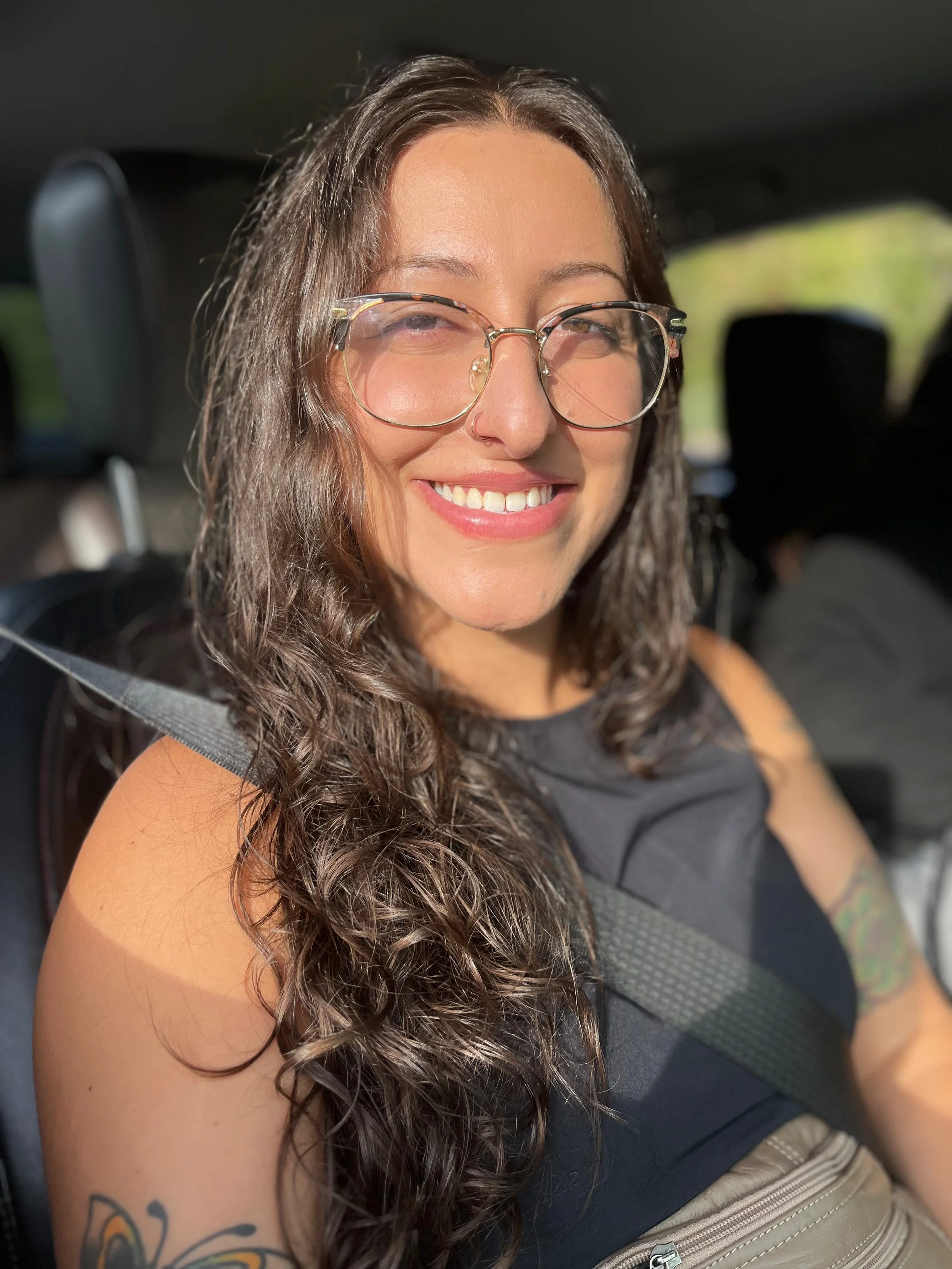Woman with long, curly hair smiling, wearing glasses and a black sleeveless top, sitting in a car with sunlight on her face.