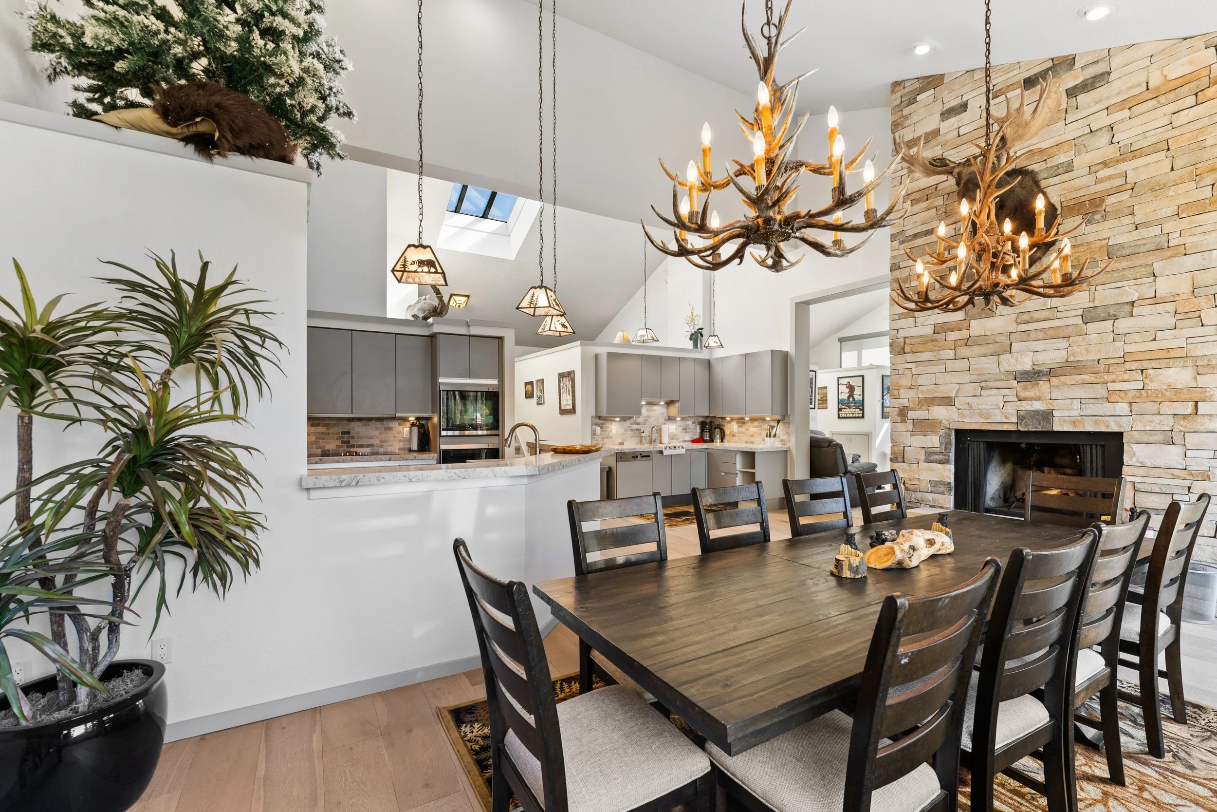 Dining room with a large wooden table and nine matching chairs, a stone fireplace, several antler chandeliers, kitchen with gray cabinets, and a potted plant in the corner.