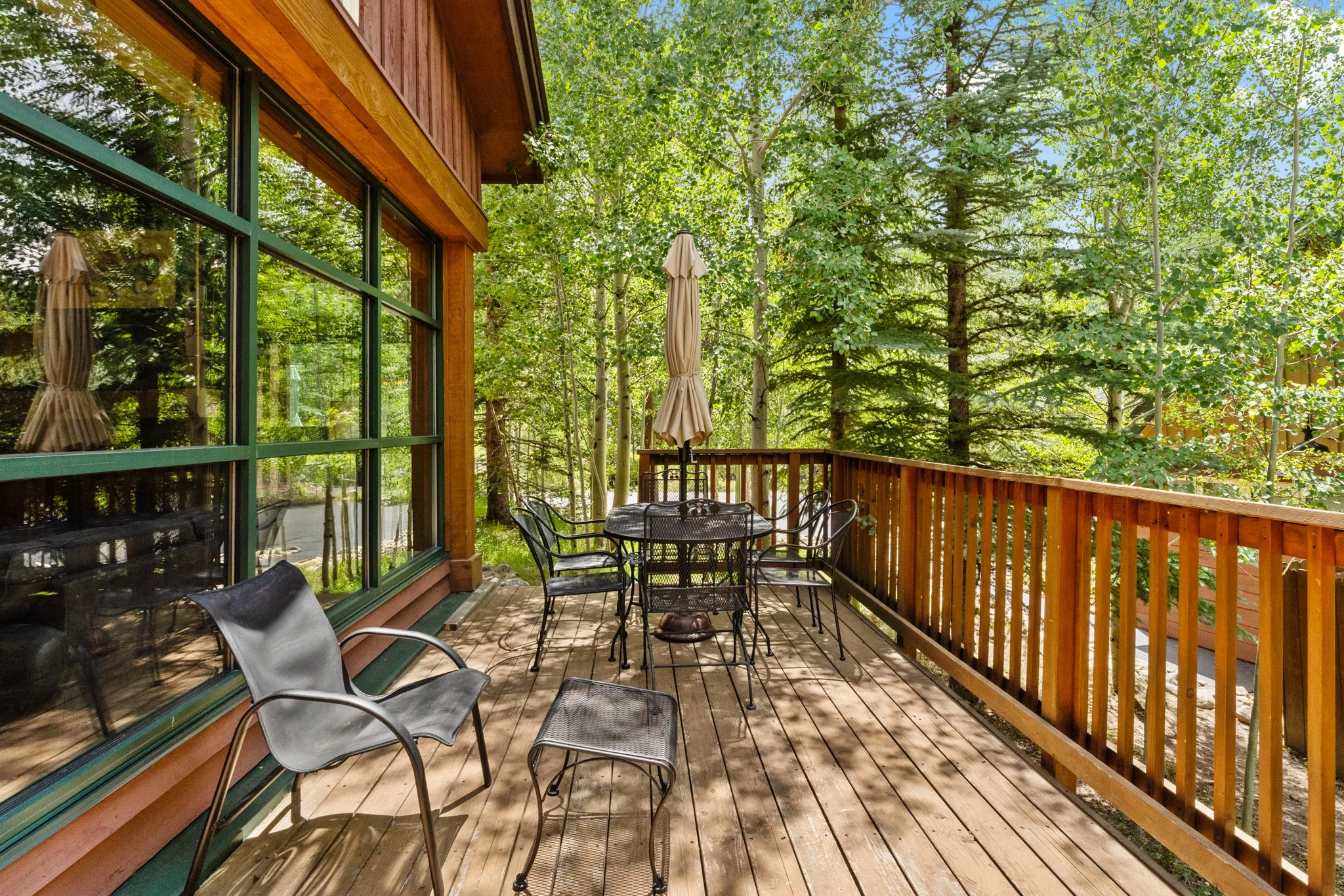 Wooden deck with outdoor furniture, including a round table with chairs and an umbrella, surrounded by green trees and a wooden railing.