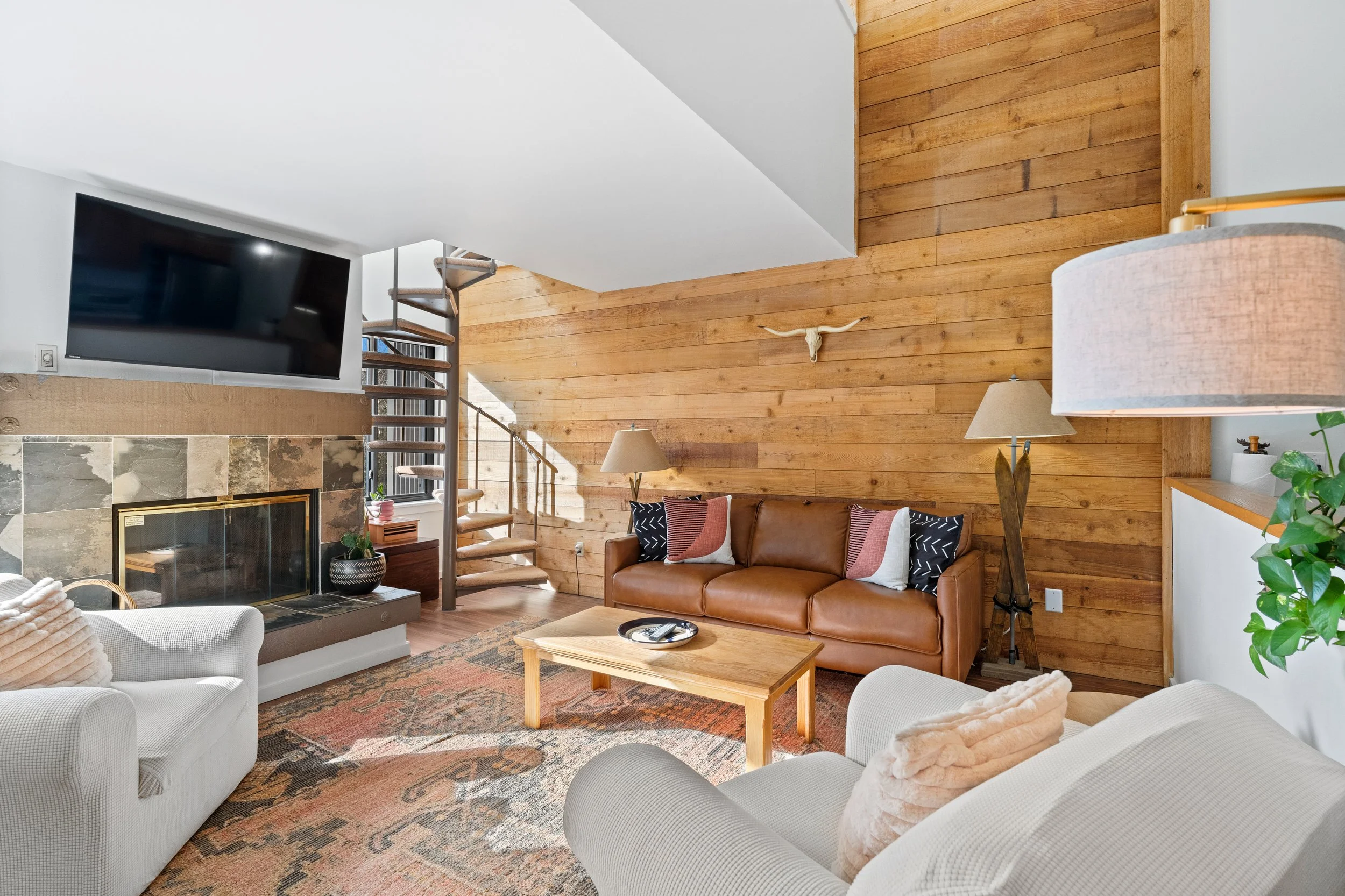 Living room with a leather sofa, two armchairs, a wooden coffee table, a fireplace, a large wall-mounted TV, a decorative bull skull, and a spiral staircase, with wooden and white walls and a patterned rug.