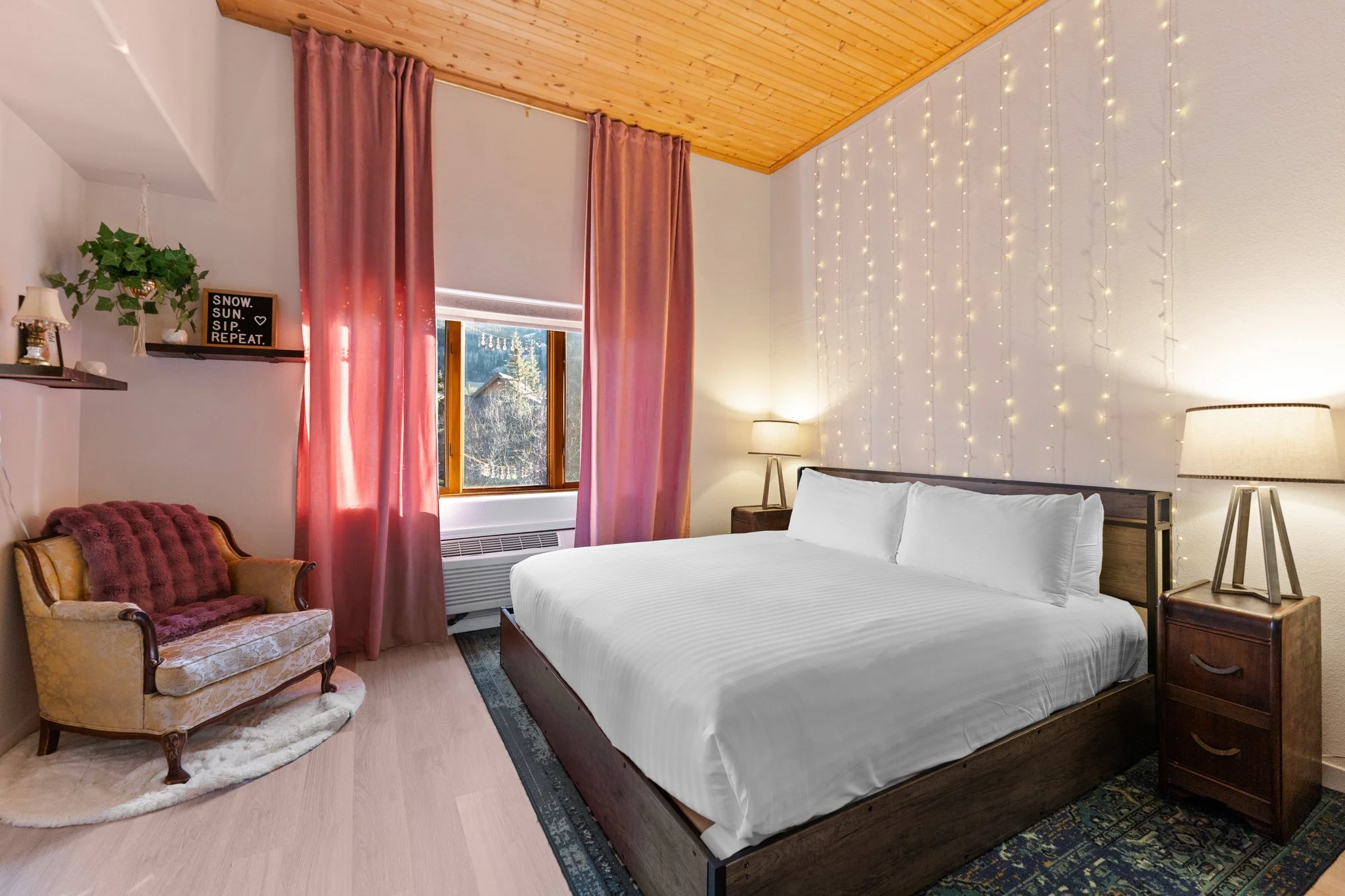 Cozy bedroom with a large bed, string lights on the wall, pink curtains, nightstands with lamps, a vintage armchair with a pink cushion, and a window showing trees outside.