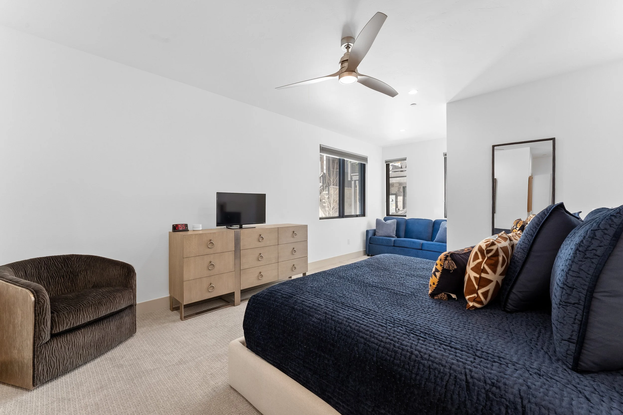 A minimalist bedroom with a large bed covered in black bedding and pillows, a small patterned armchair, a wooden dresser with a TV, a blue sofa, and a ceiling fan.