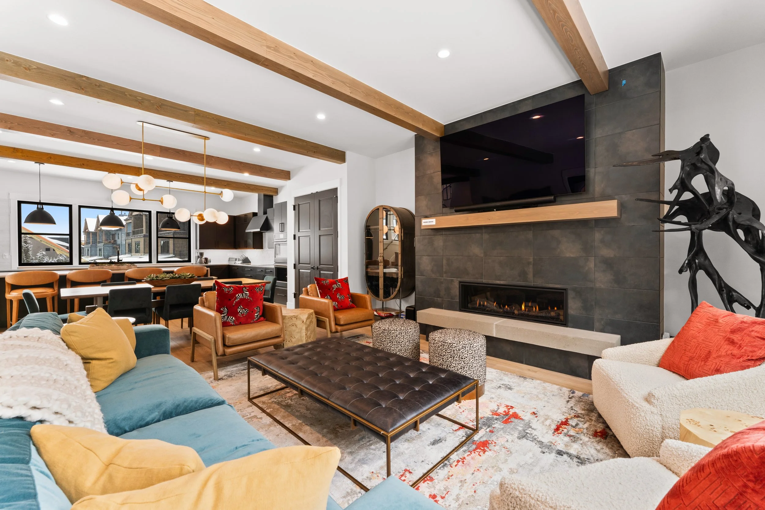 Modern living room with multicolored furniture, a fireplace, a large flat-screen TV on a black tiled wall, and a dining area in the background with black chandeliers and windows showing a snowy street view.