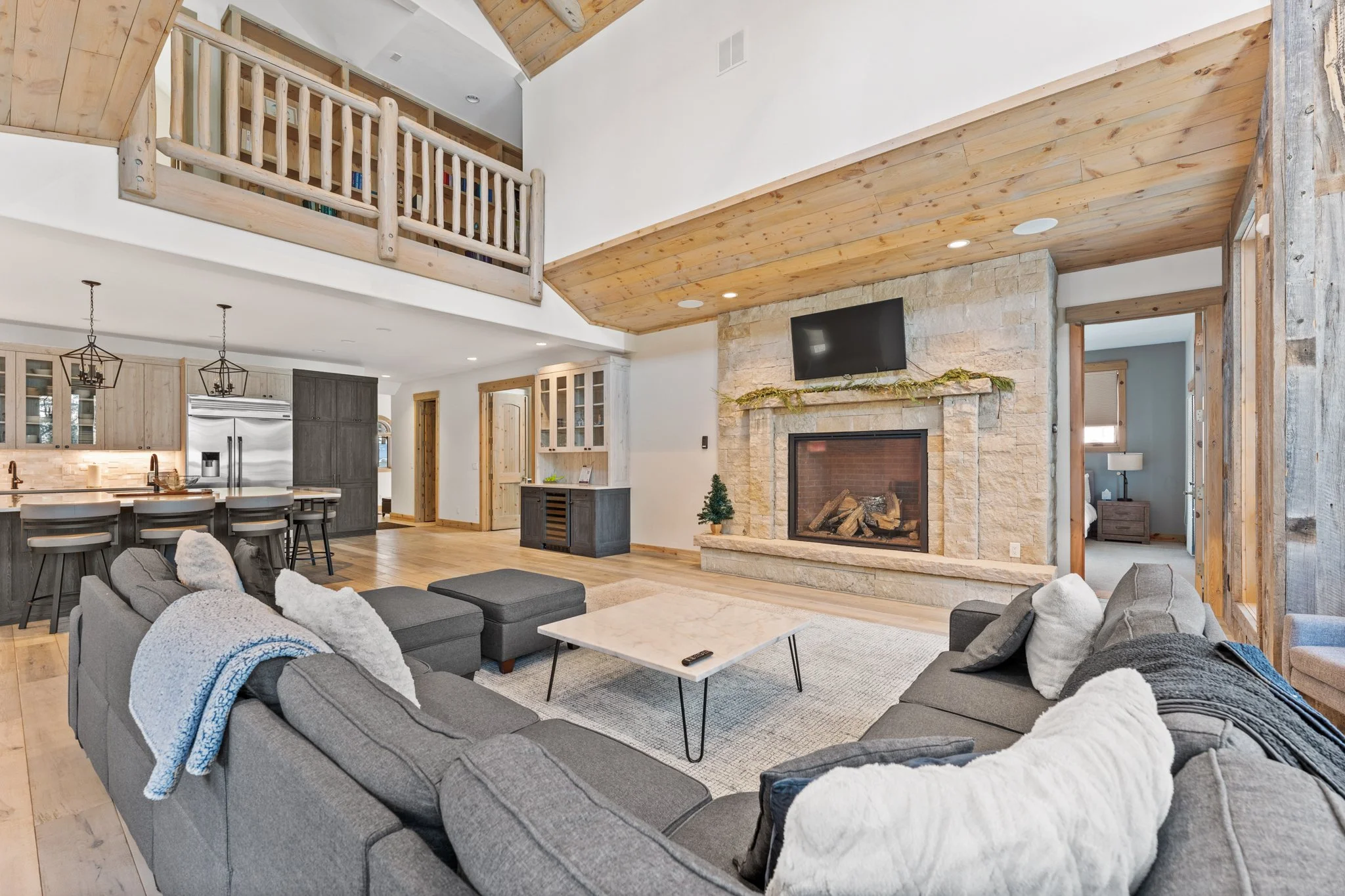 A spacious living room with modern decor featuring a large gray sectional sofa with white pillows, a fireplace with a TV mounted above, a wooden mantel decorated with greenery, and a small Christmas tree. The room has wood accents, a vaulted ceiling, and an open layout connecting to the kitchen and a bedroom visible in the background.