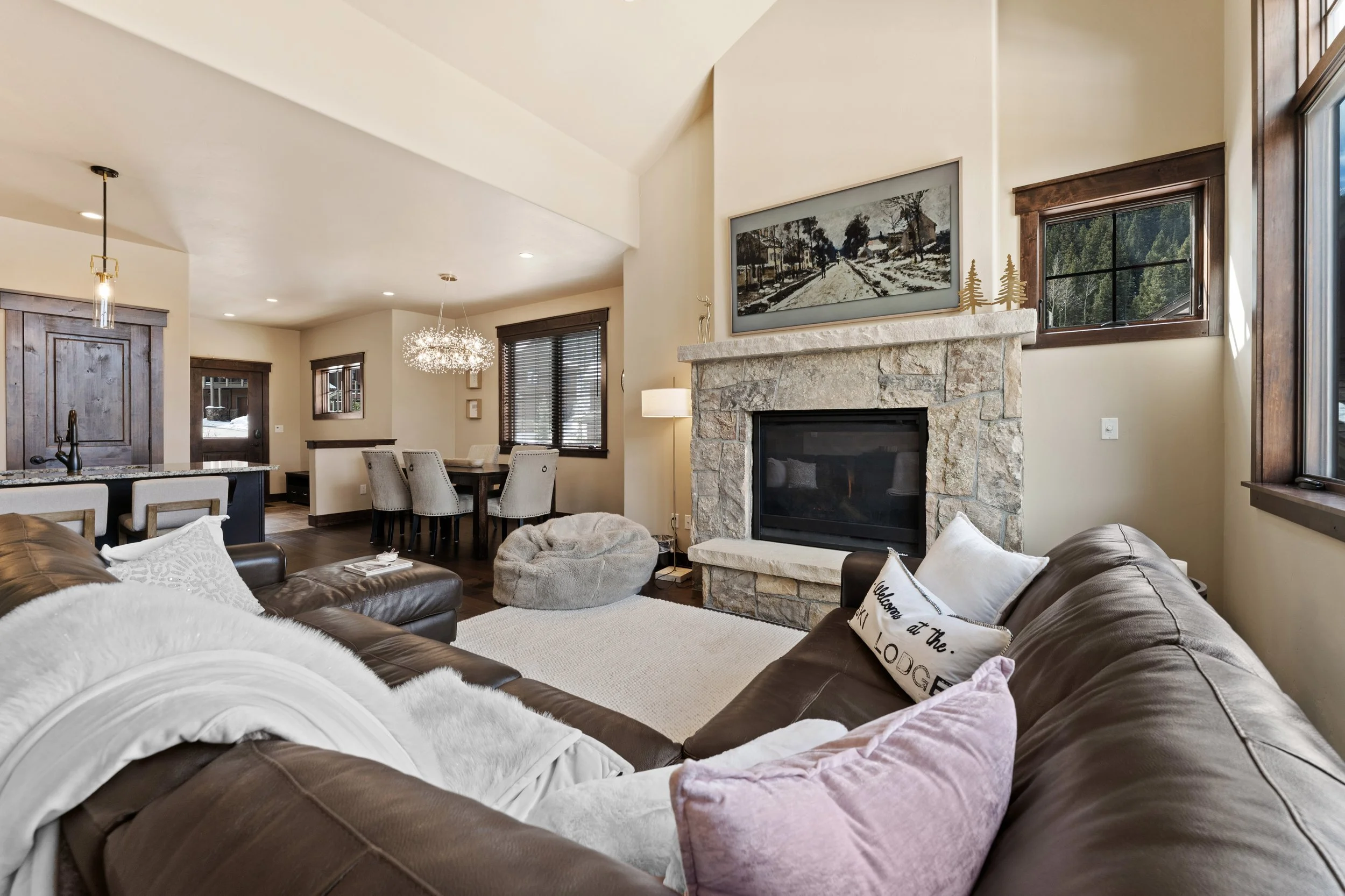 Living room with a stone fireplace, brown leather sofas, decorative pillows, large windows, and a fireplace with a painting above it.