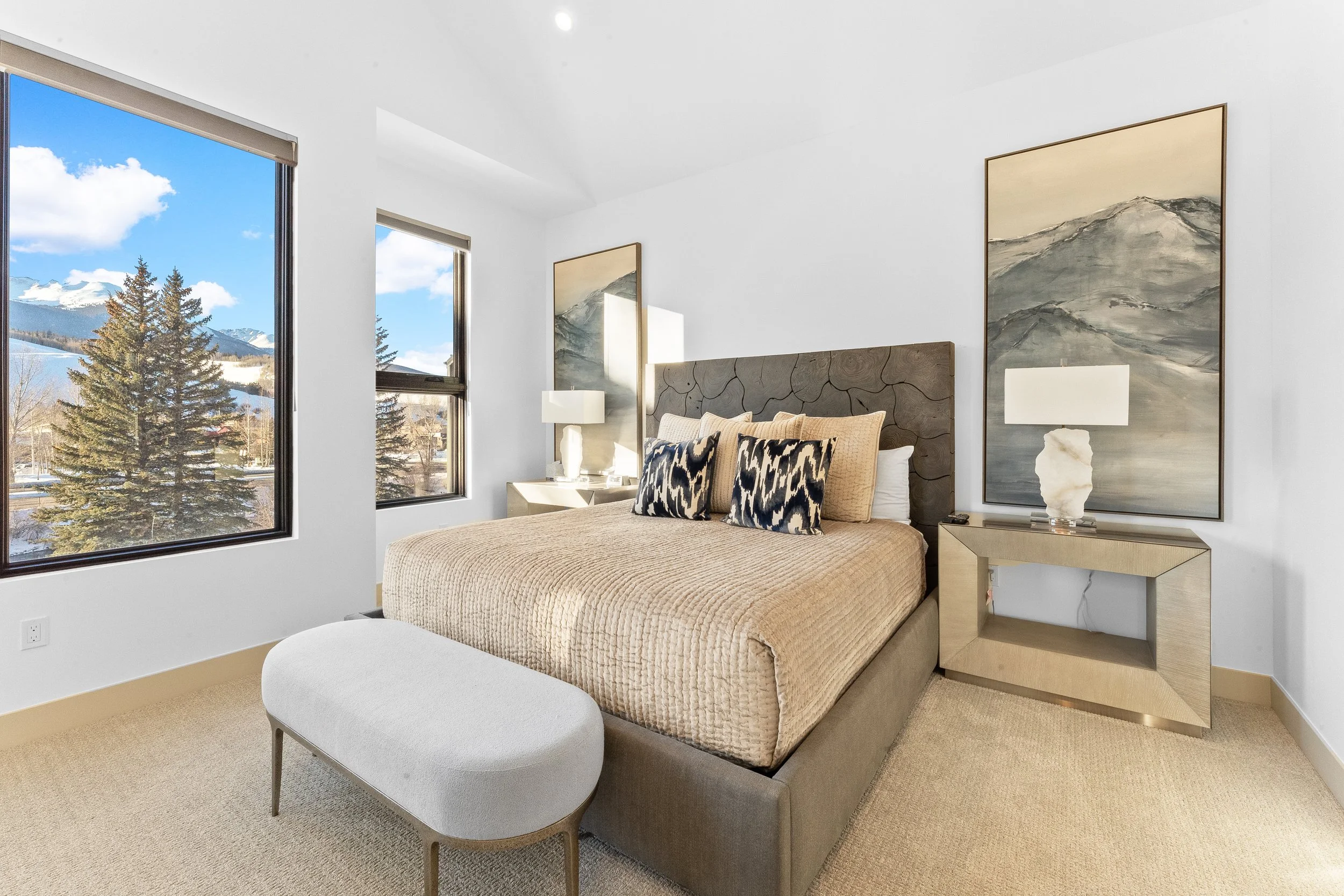 A bedroom with a large bed, beige bedding, and decorative pillows. Two nightstands with white lamps. Two large landscape paintings behind the nightstands. Large windows showing trees and mountains outside.